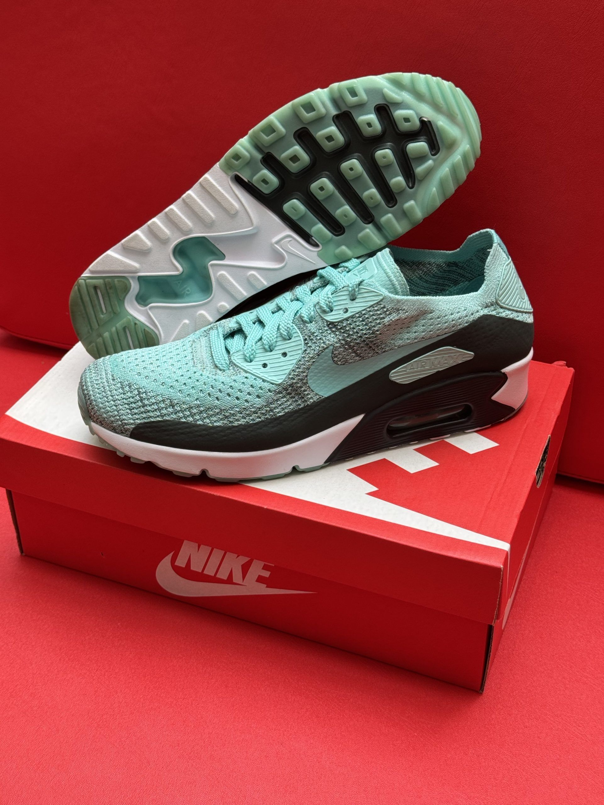 Blue Nike Air Max 90 sneakers on a red Nike box, one shoe upside down showing the green sole.