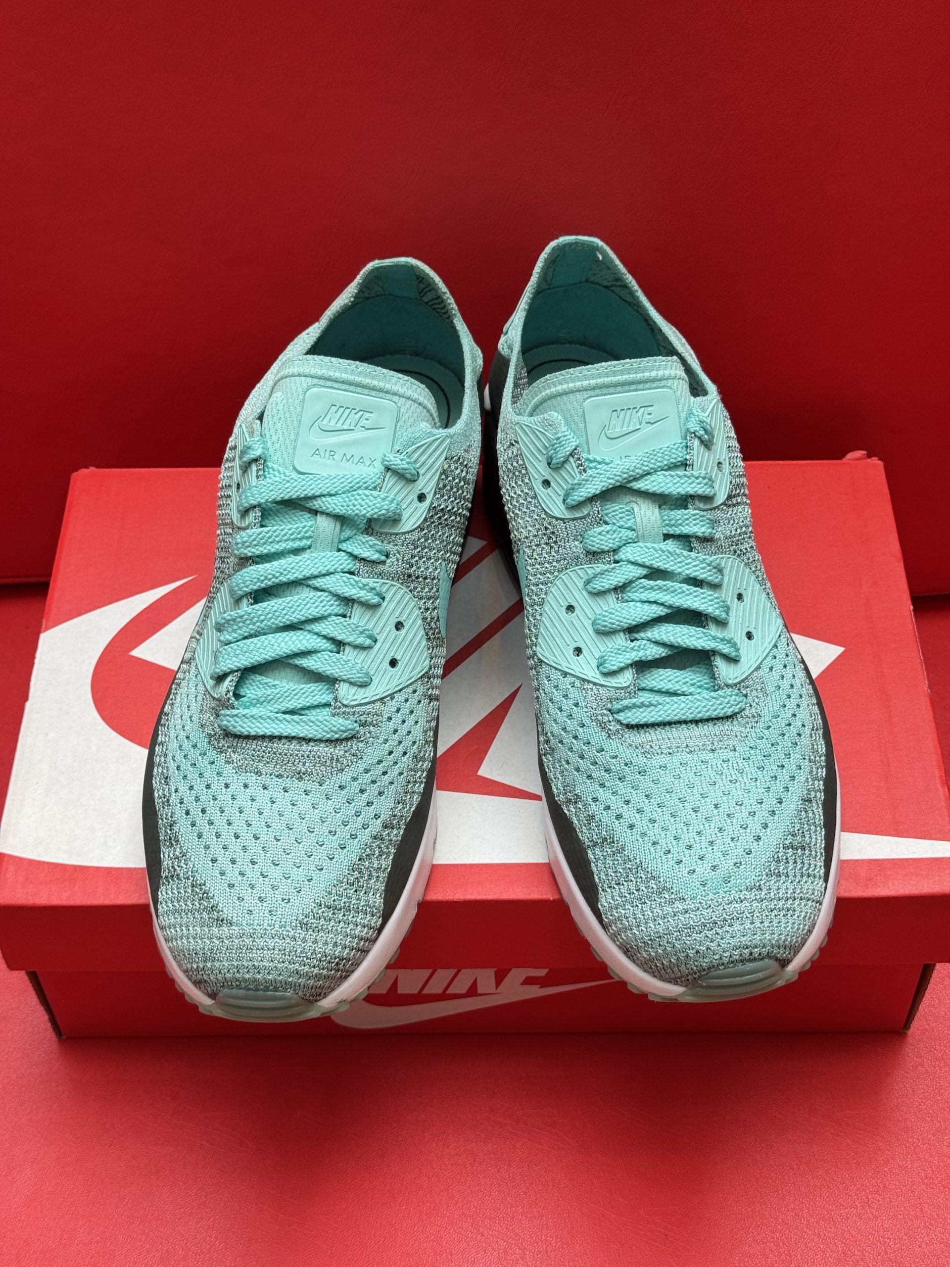 Blue Nike Air Max 90 sneakers on a red Nike box, one shoe upside down showing the green sole.