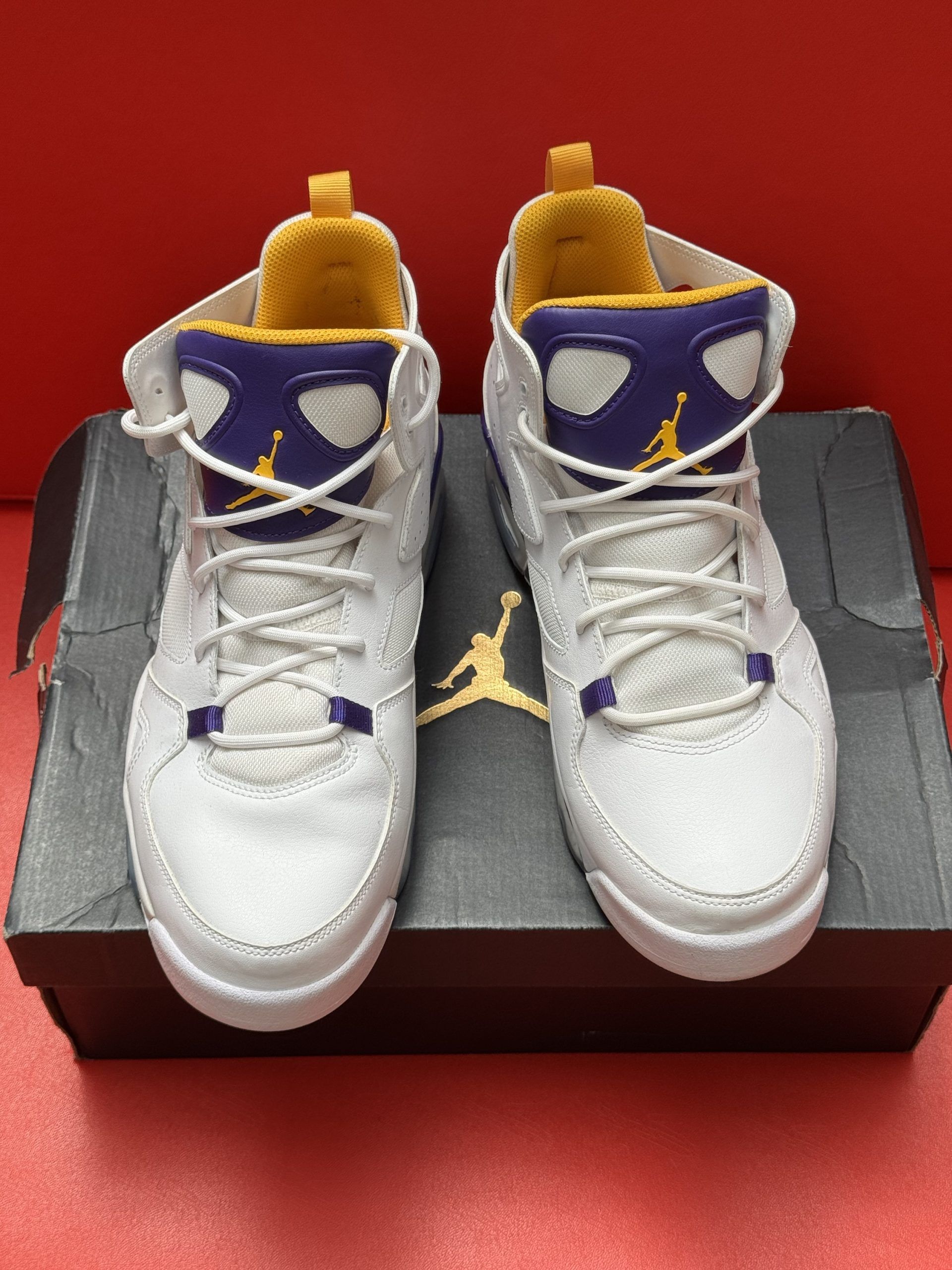 White and purple Air Jordan sneakers with yellow accents, atop a shoebox, on a red surface.