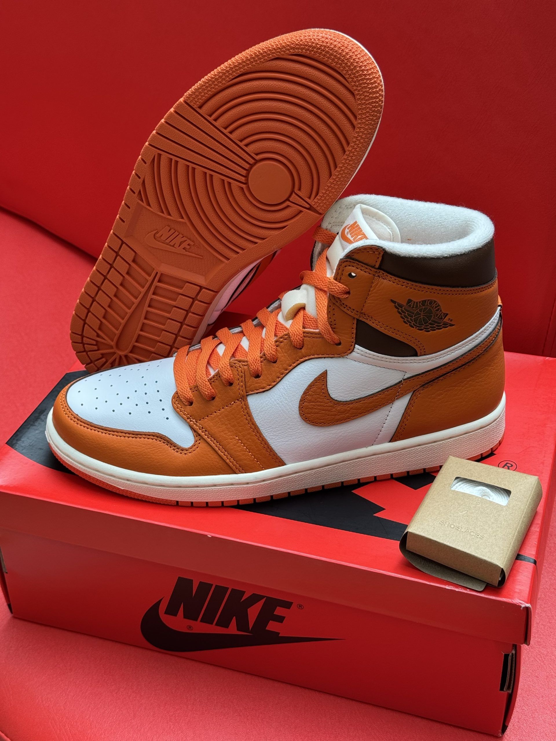 Orange and white Air Jordan 1 sneaker, box, and shoe sole on a red surface.
