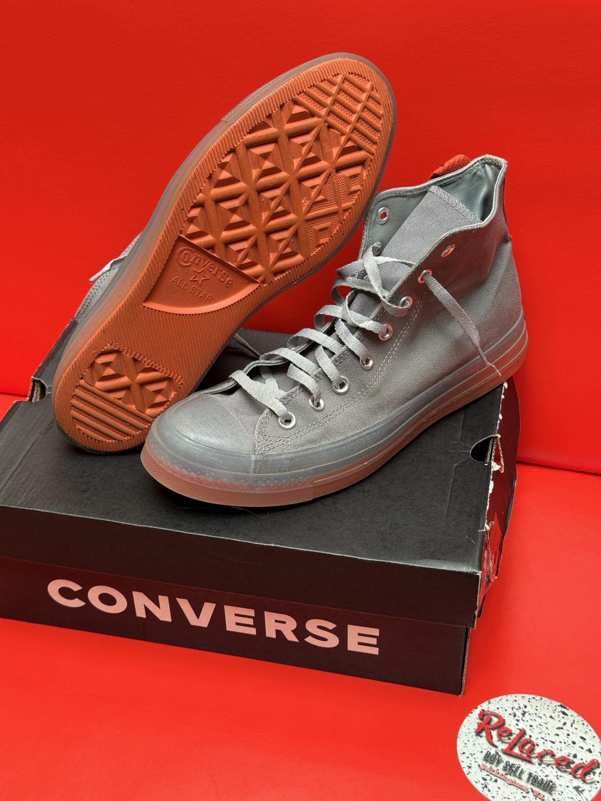 Gray Converse high-top sneakers with a brown sole, sitting on a black Converse box against a red background.