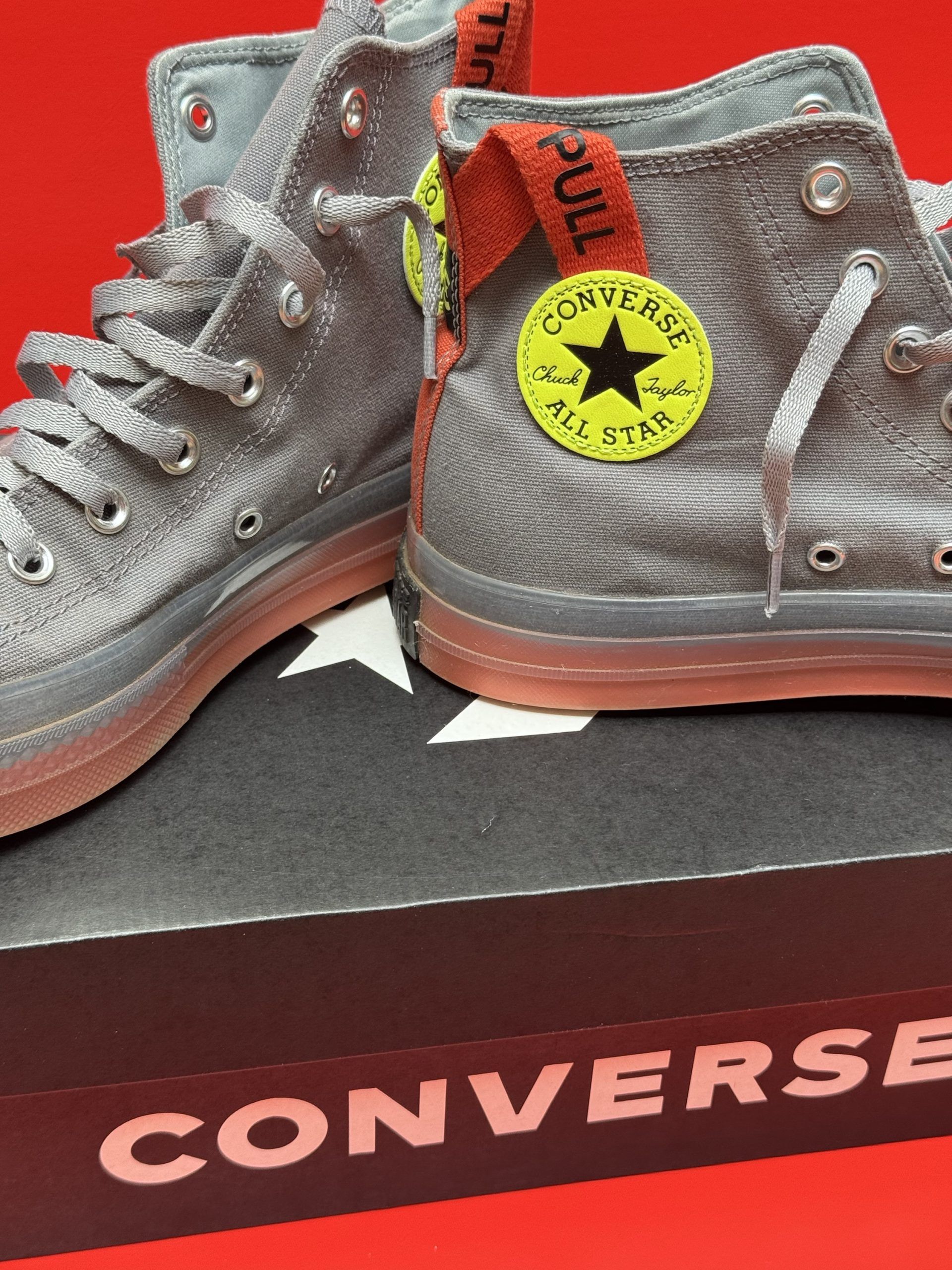 Gray Converse high-top sneakers with a brown sole, sitting on a black Converse box against a red background.