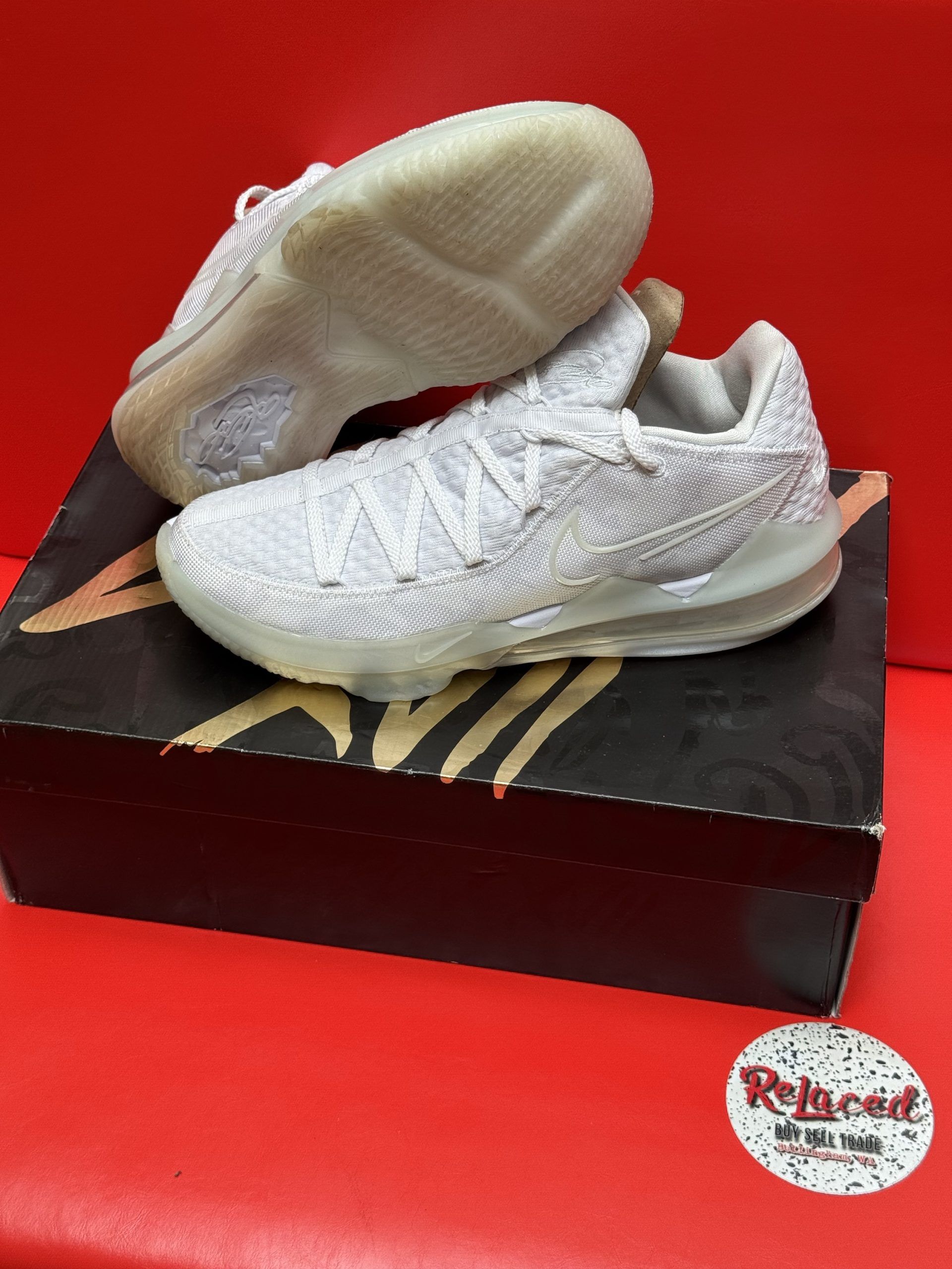 White sneakers on a box, red background. Sneakers are low-cut, textured, and have a translucent sole. Box is dark with gold text.