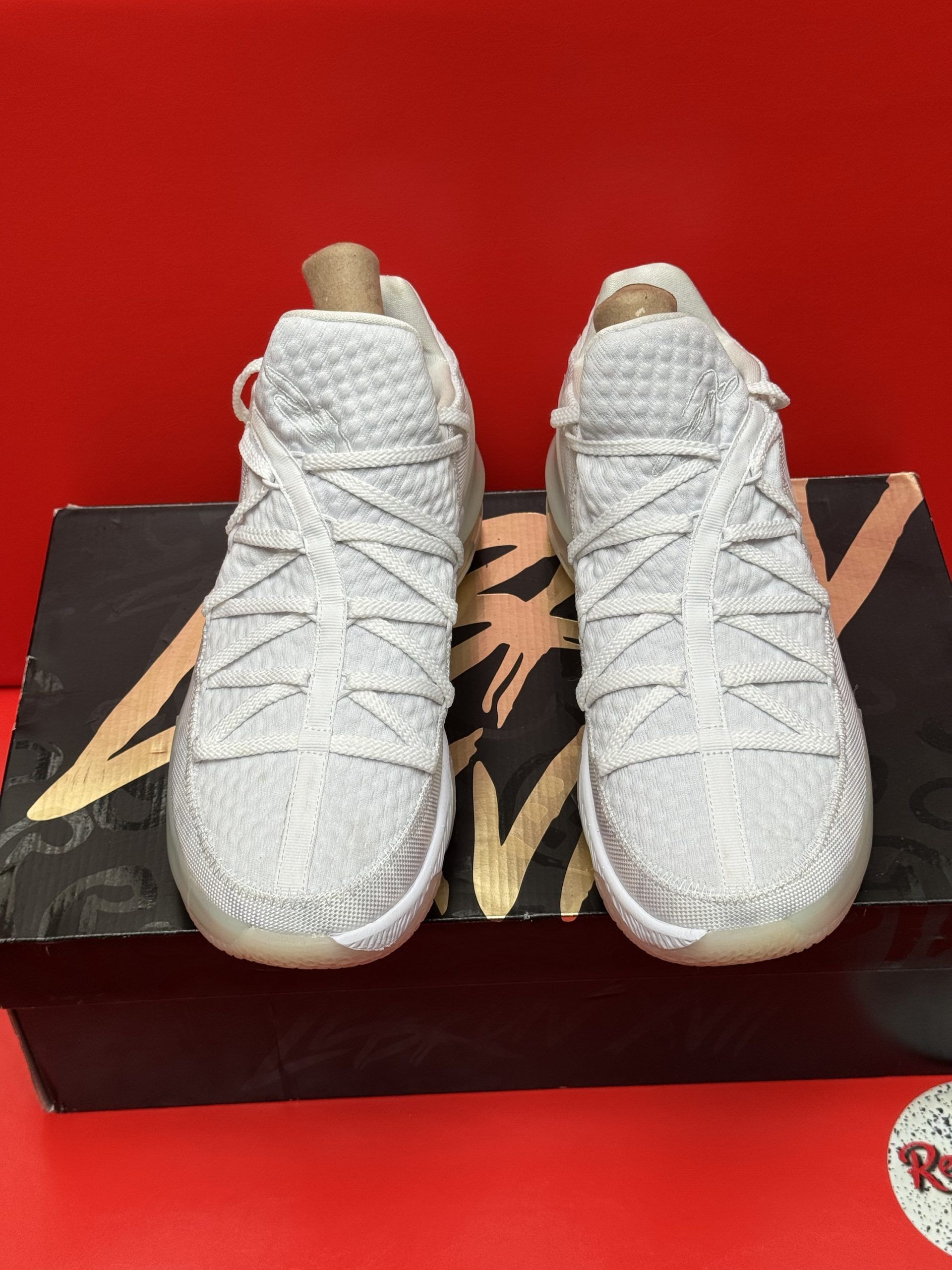 White sneakers on a box, red background. Sneakers are low-cut, textured, and have a translucent sole. Box is dark with gold text.
