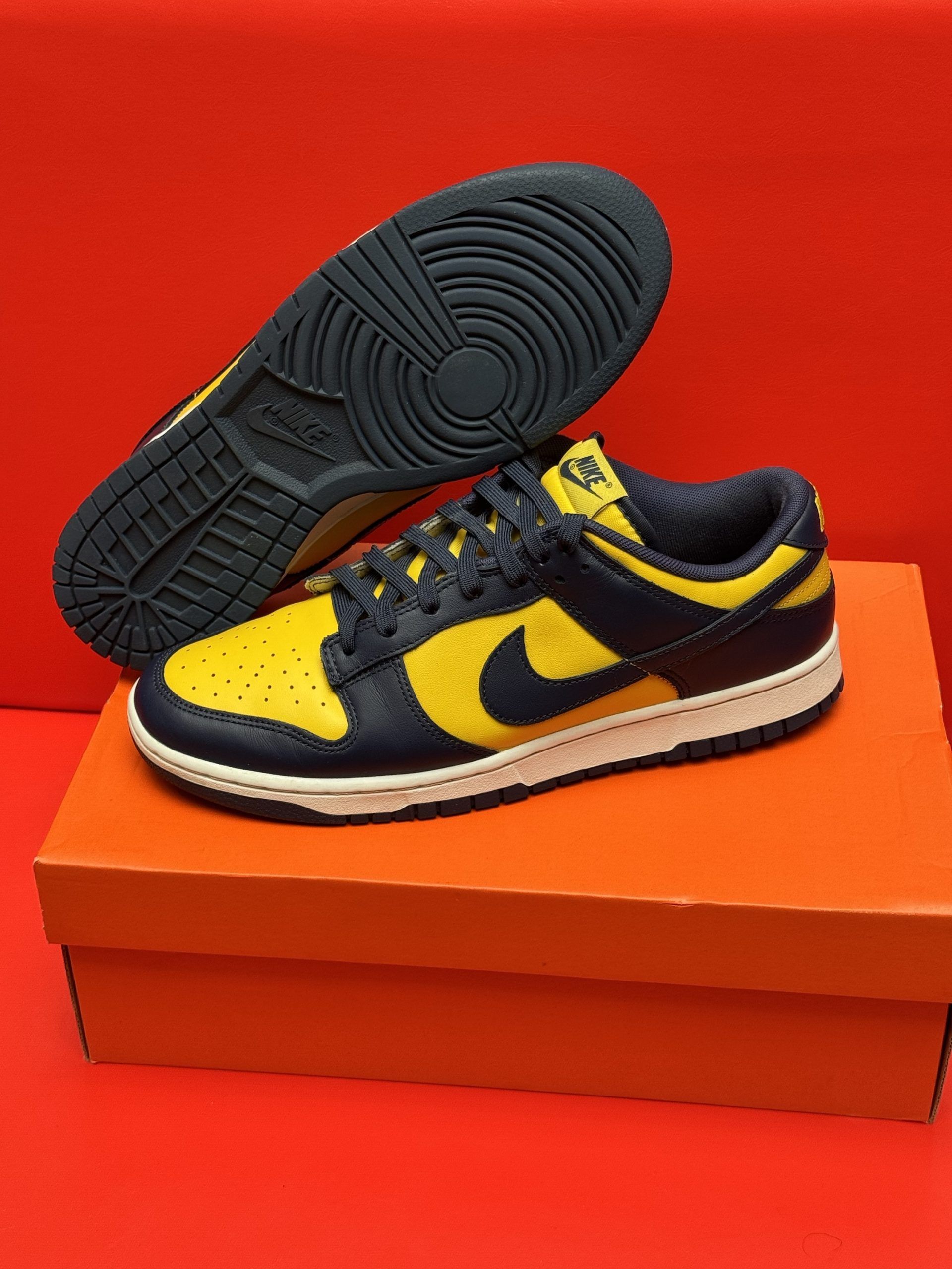 Yellow and navy Nike Dunk Low sneakers, box, and sole displayed against a red background.