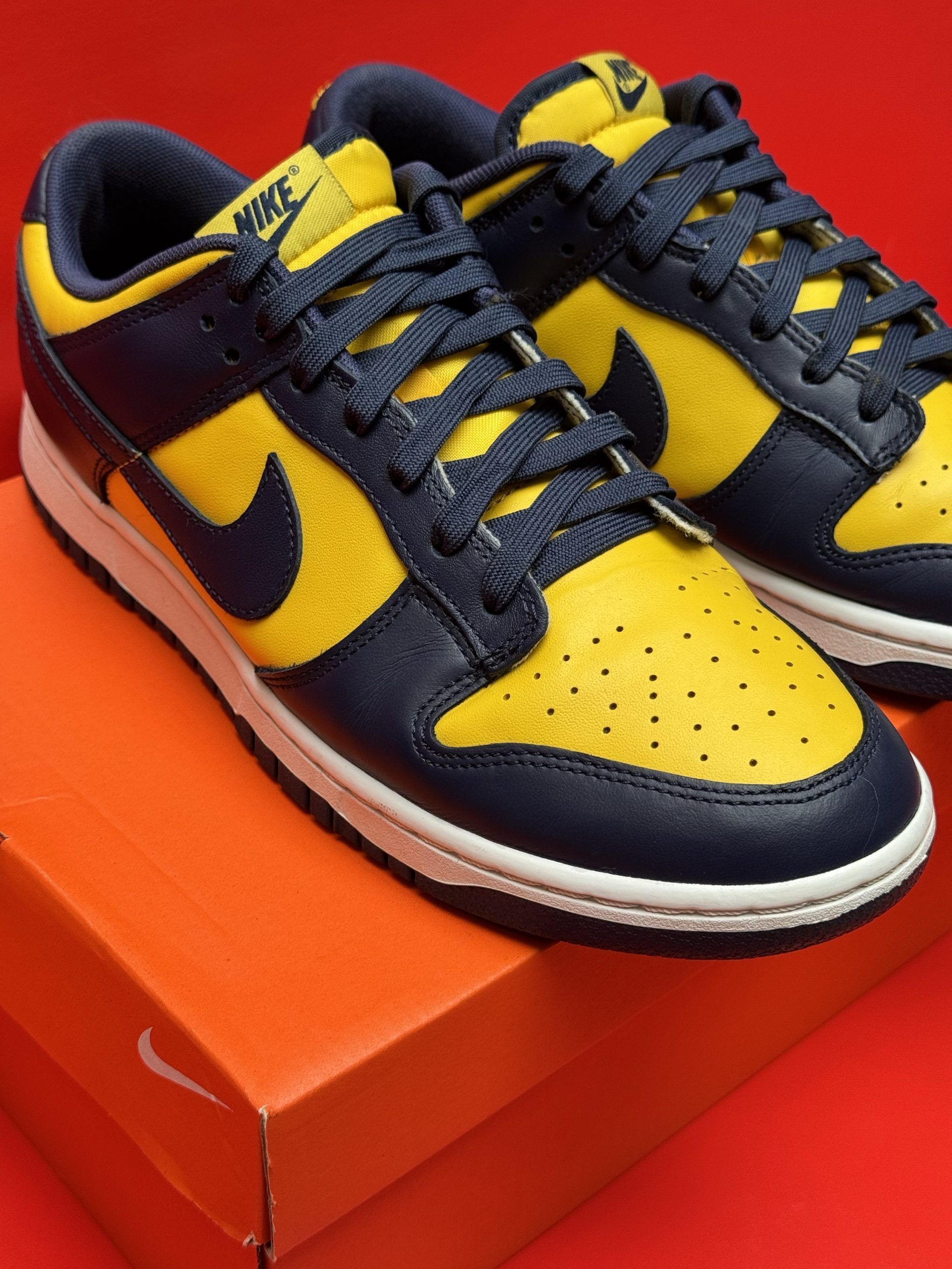 Yellow and navy Nike Dunk Low sneakers, box, and sole displayed against a red background.