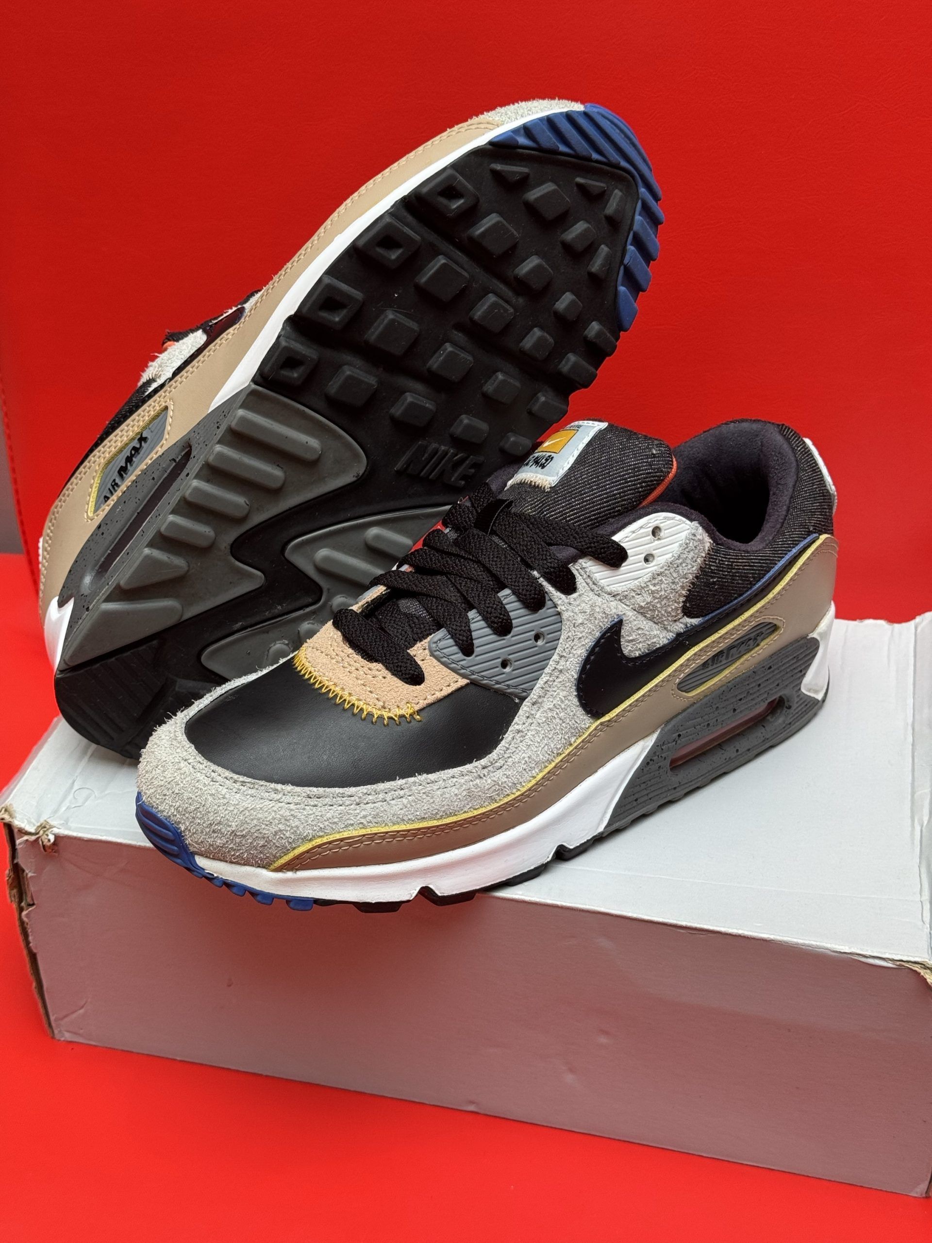 Nike Air Max 90 sneakers with black, tan, and blue accents, set on a red surface.