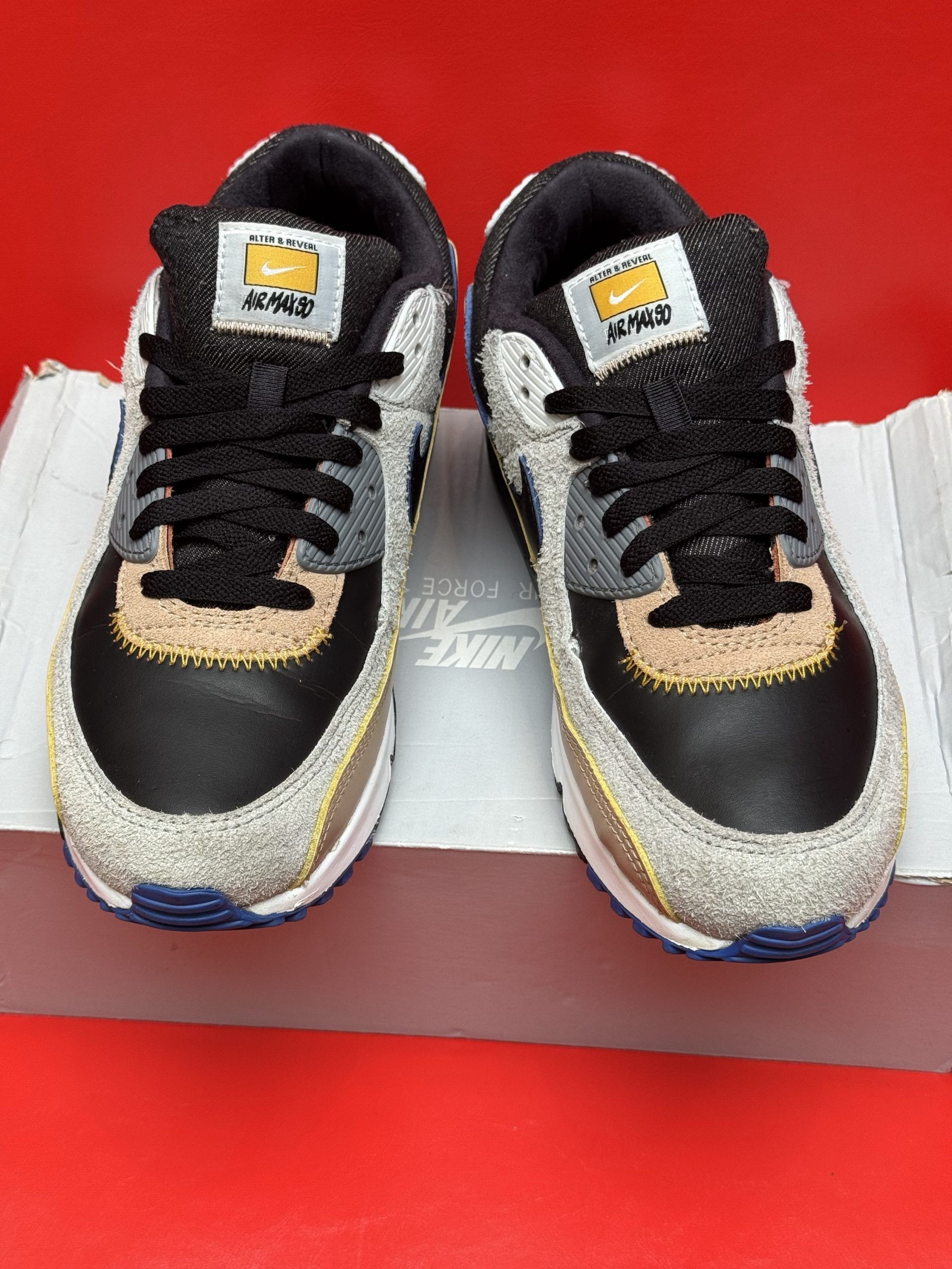 Nike Air Max 90 sneakers with black, tan, and blue accents, set on a red surface.