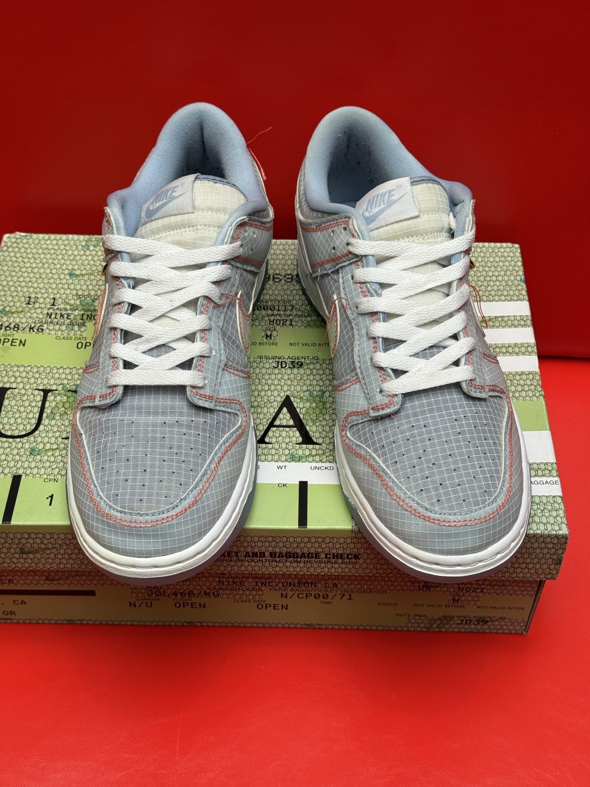 Blue and beige Nike Dunk Low sneakers with white laces on a box, red background.