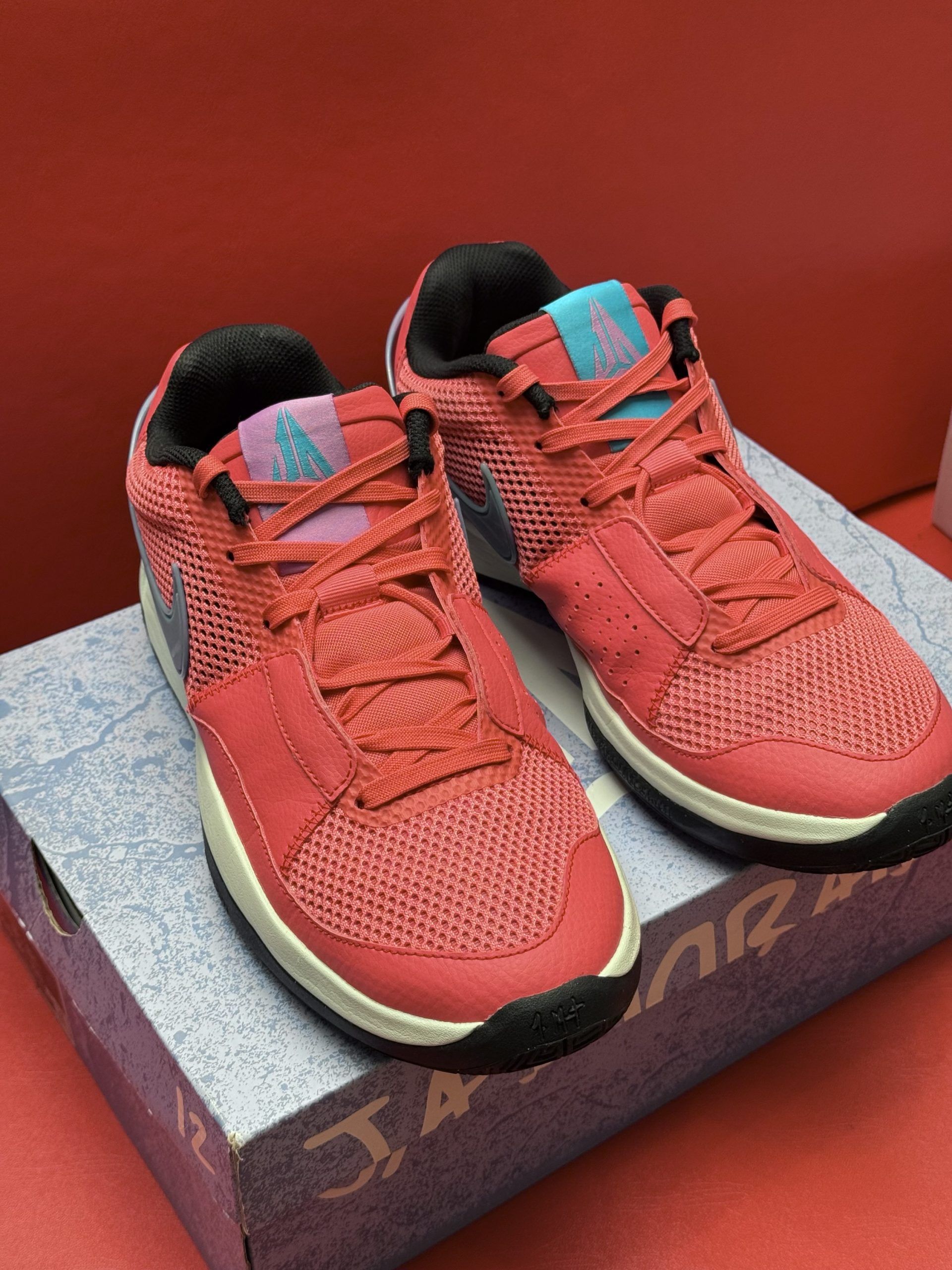 Red and blue Nike Ja Morant sneakers on a purple box, one shoe up, resting on a red surface.