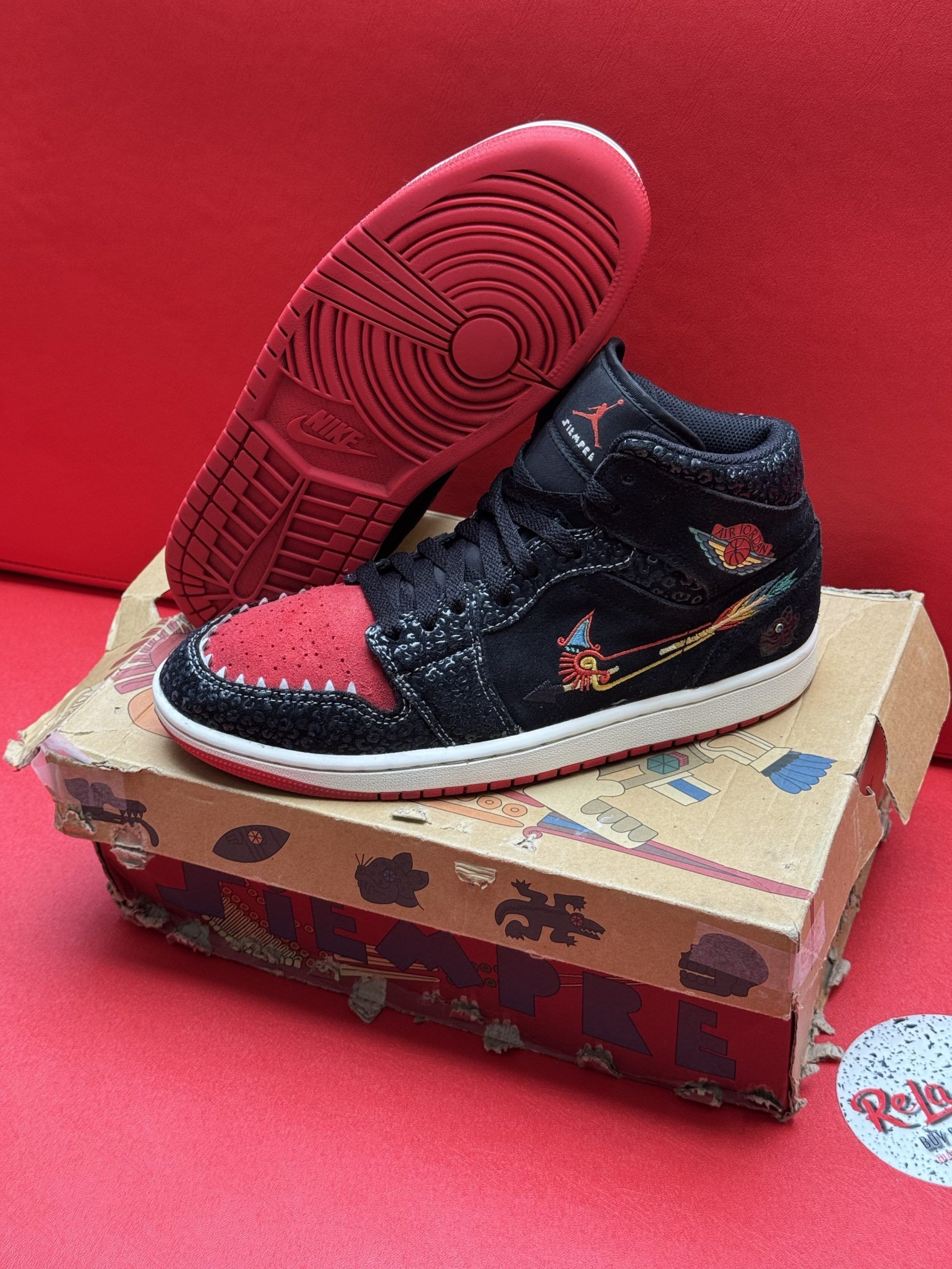 Black and red Air Jordan sneakers on a cardboard box, sole up, resting on a red surface.
