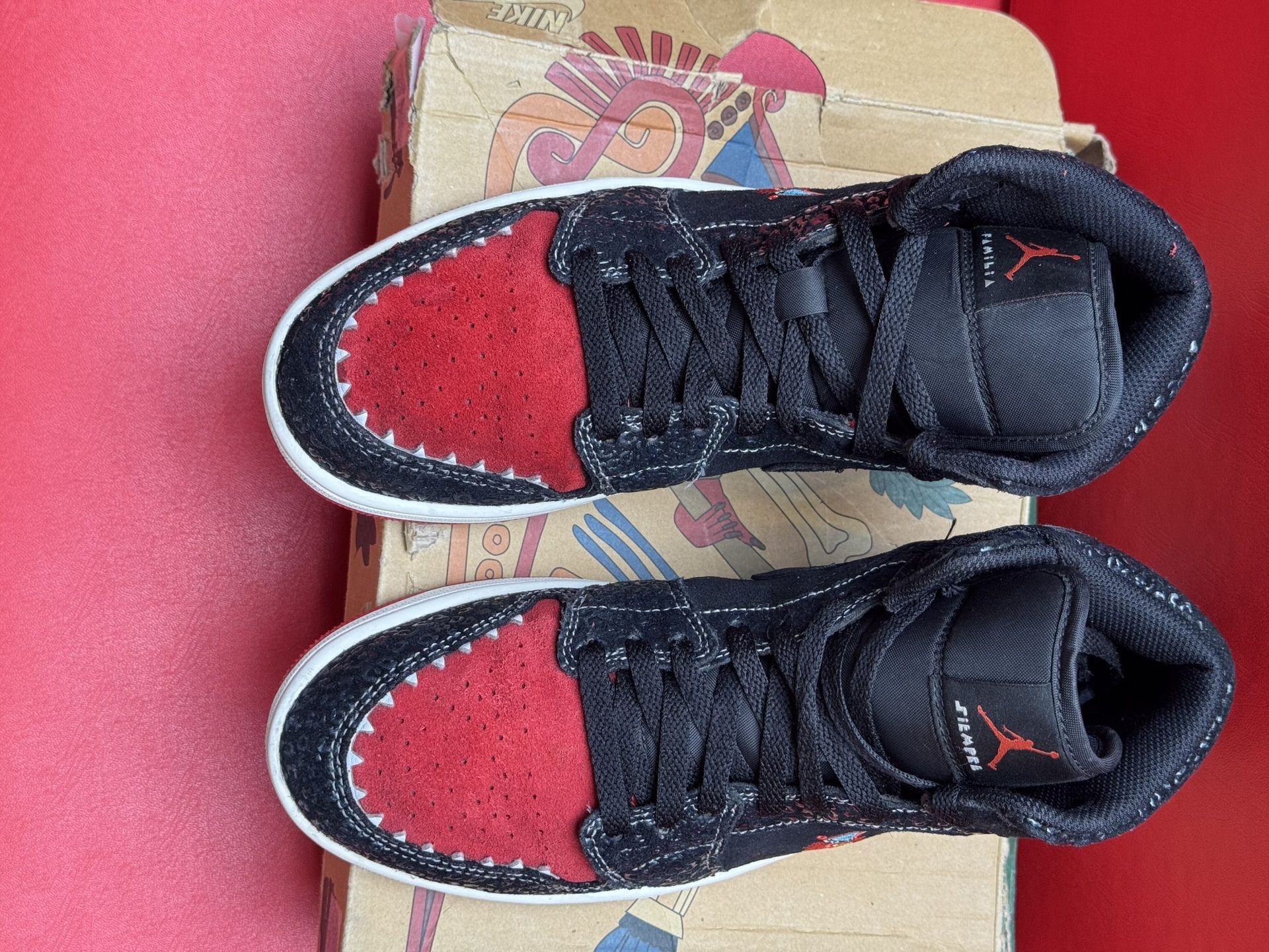 Black and red Air Jordan sneakers on a cardboard box, sole up, resting on a red surface.