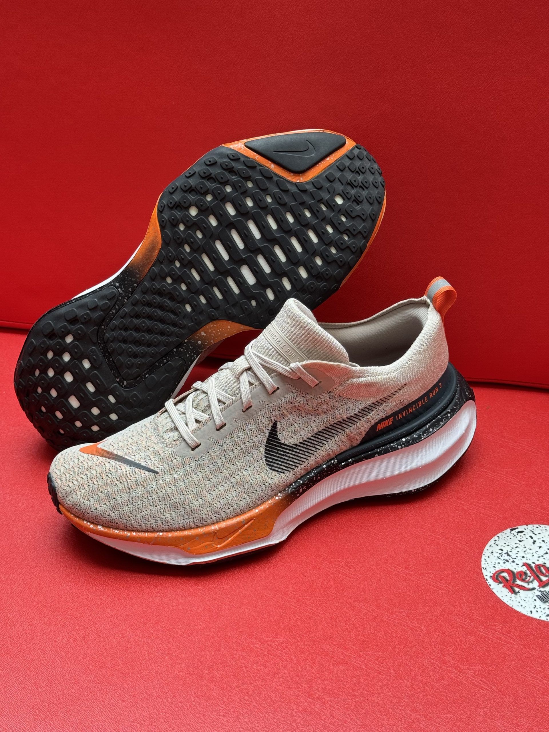 Beige Nike running shoe with orange accents, black sole, resting on a red surface.