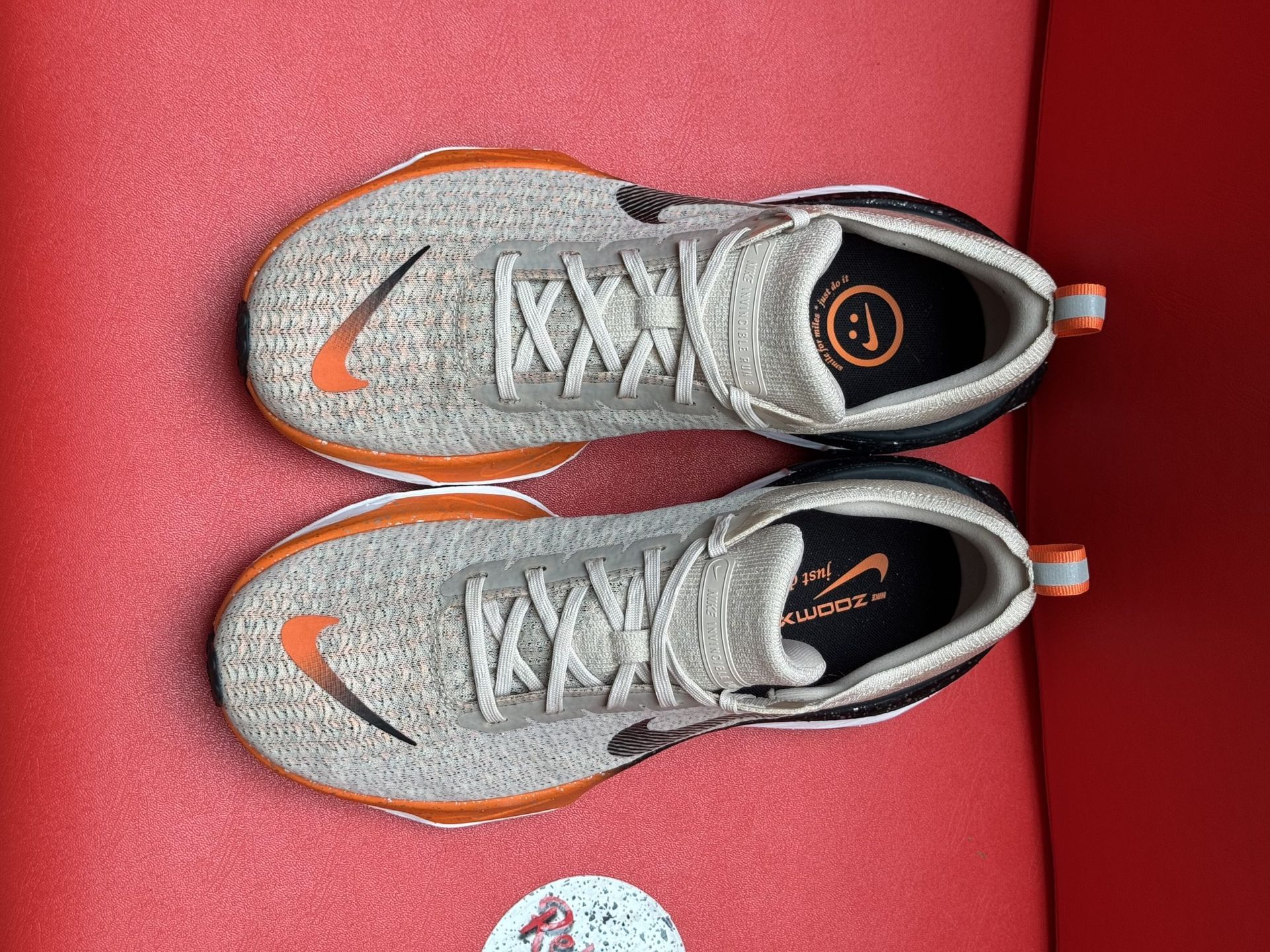 Beige Nike running shoe with orange accents, black sole, resting on a red surface.