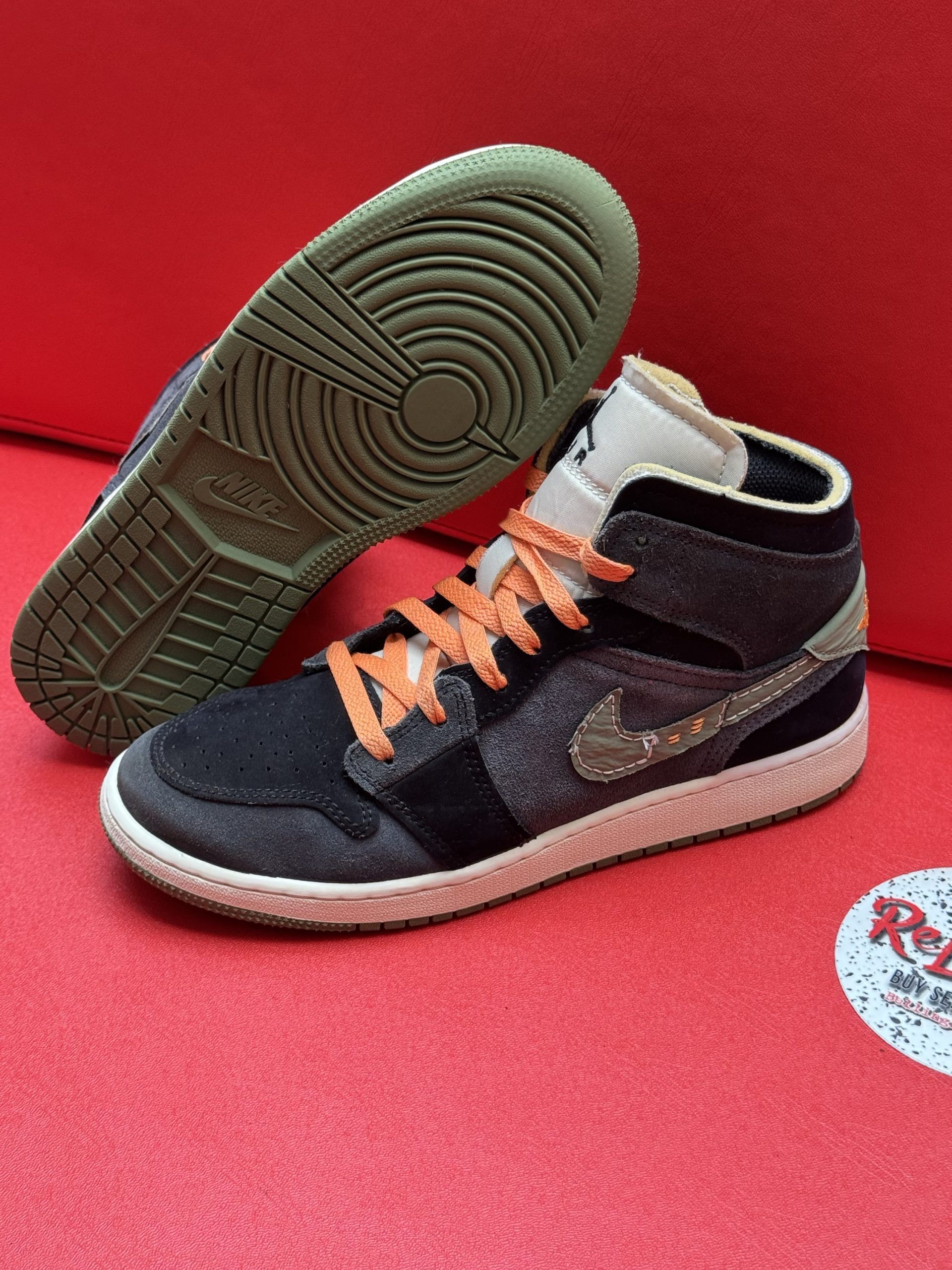 Black, gray, and orange Nike Air Jordan 1 mid sneakers on a red surface, one sole up.