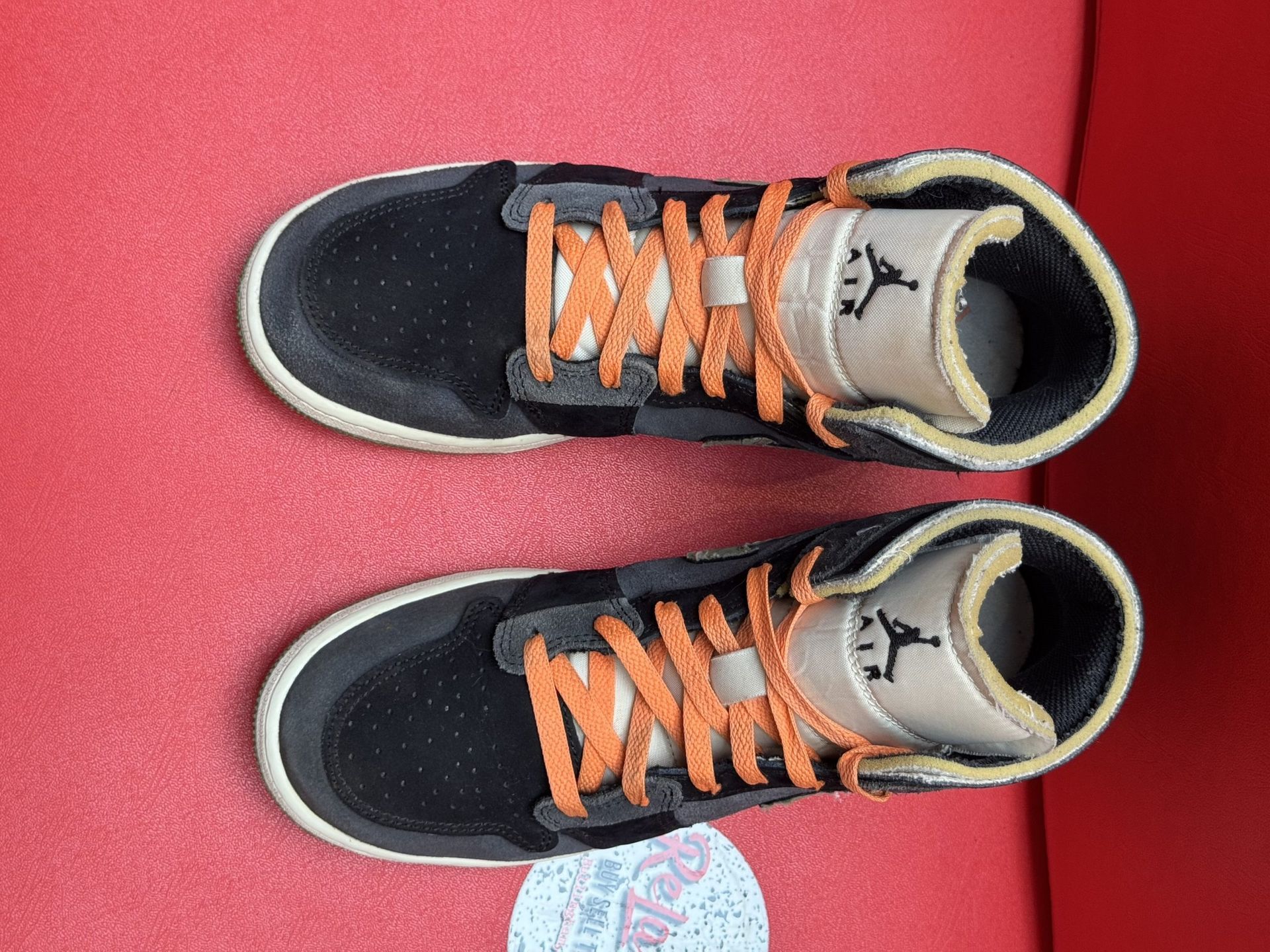 Black, gray, and orange Nike Air Jordan 1 mid sneakers on a red surface, one sole up.