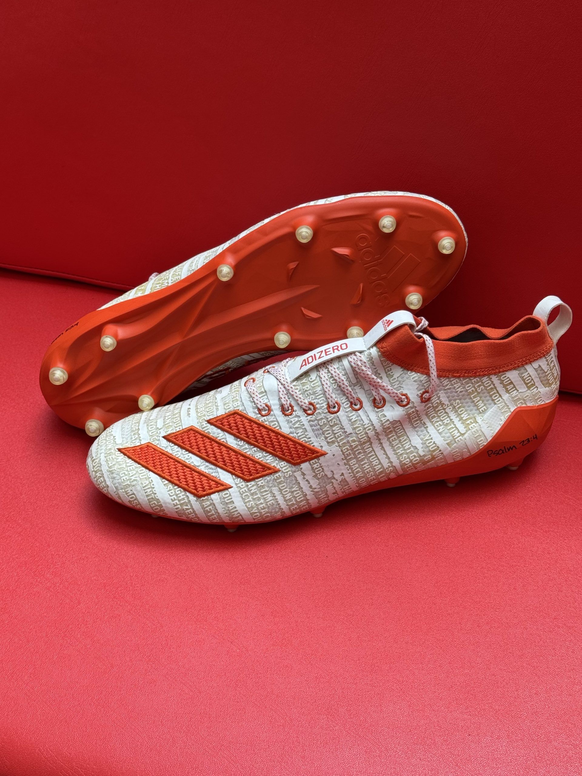 White and orange Adidas football cleats on a red surface, showcasing the sole with cleats and the shoe's design.