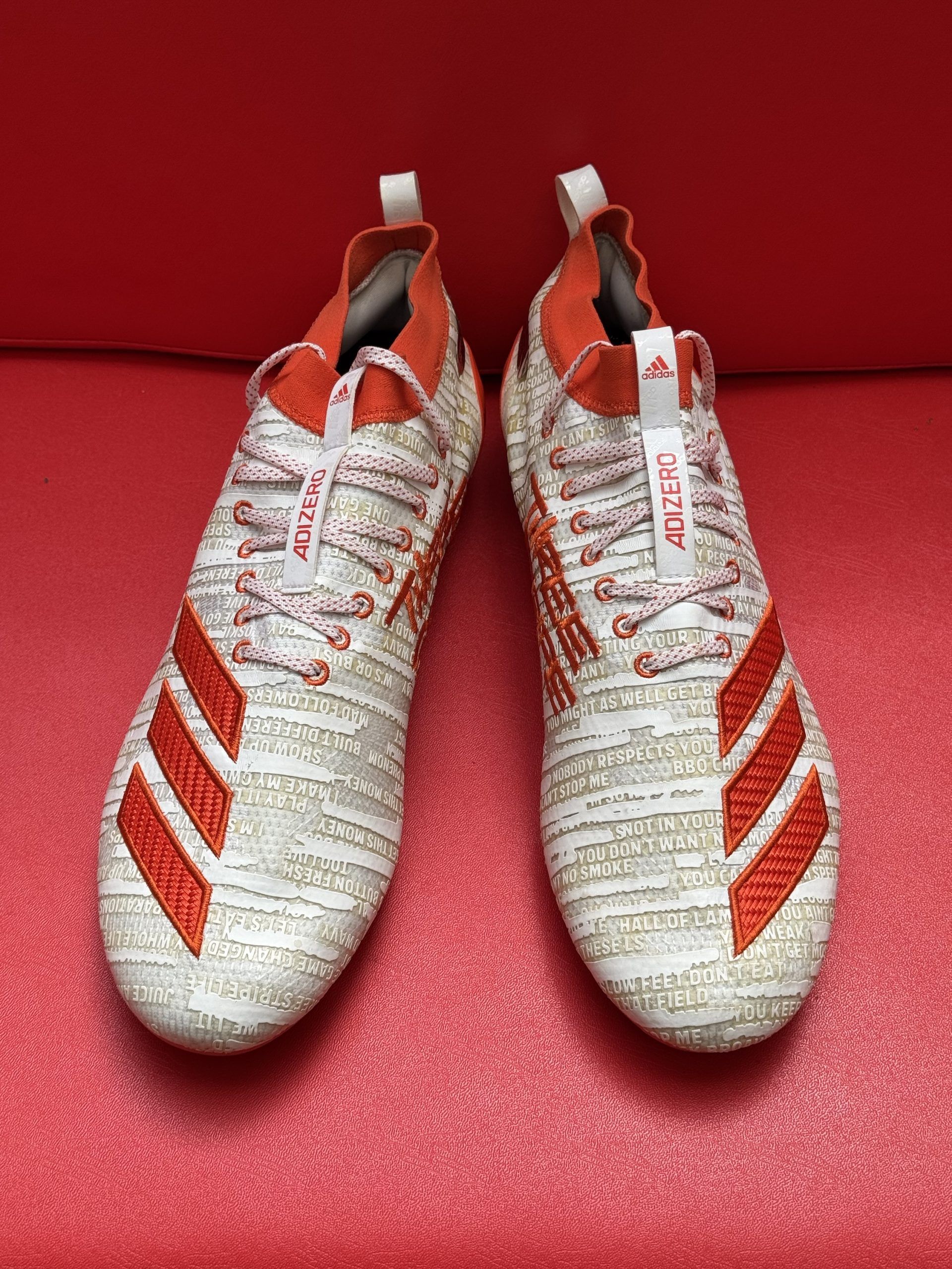 White and orange Adidas football cleats on a red surface, showcasing the sole with cleats and the shoe's design.