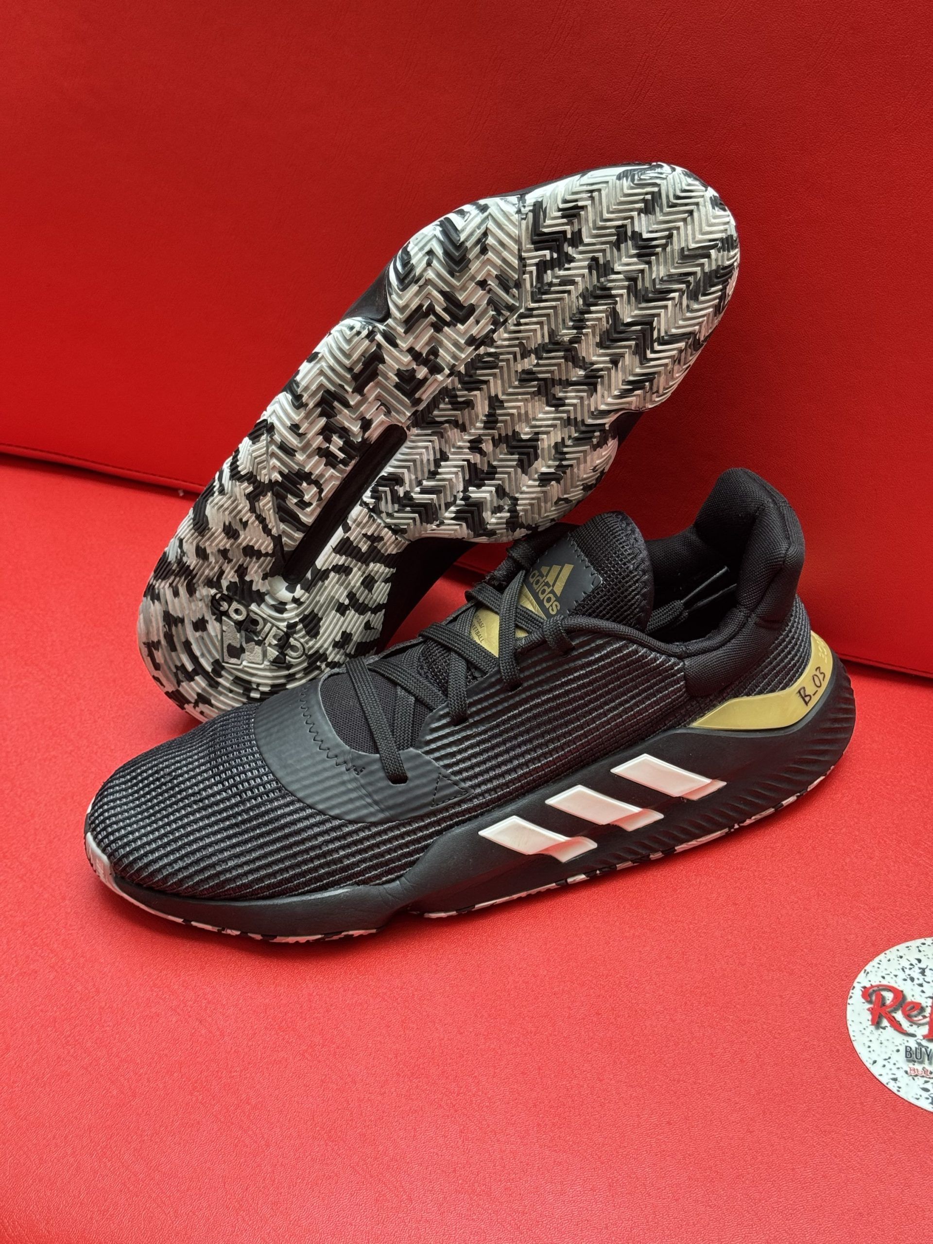 Black and gold Adidas basketball shoes with white stripes on a red surface.