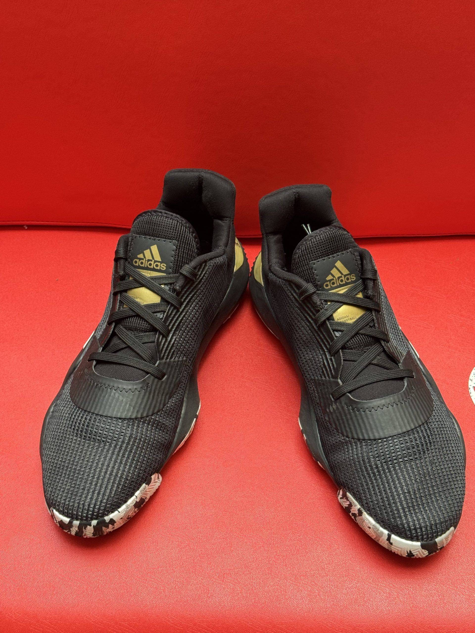 Black and gold Adidas basketball shoes with white stripes on a red surface.