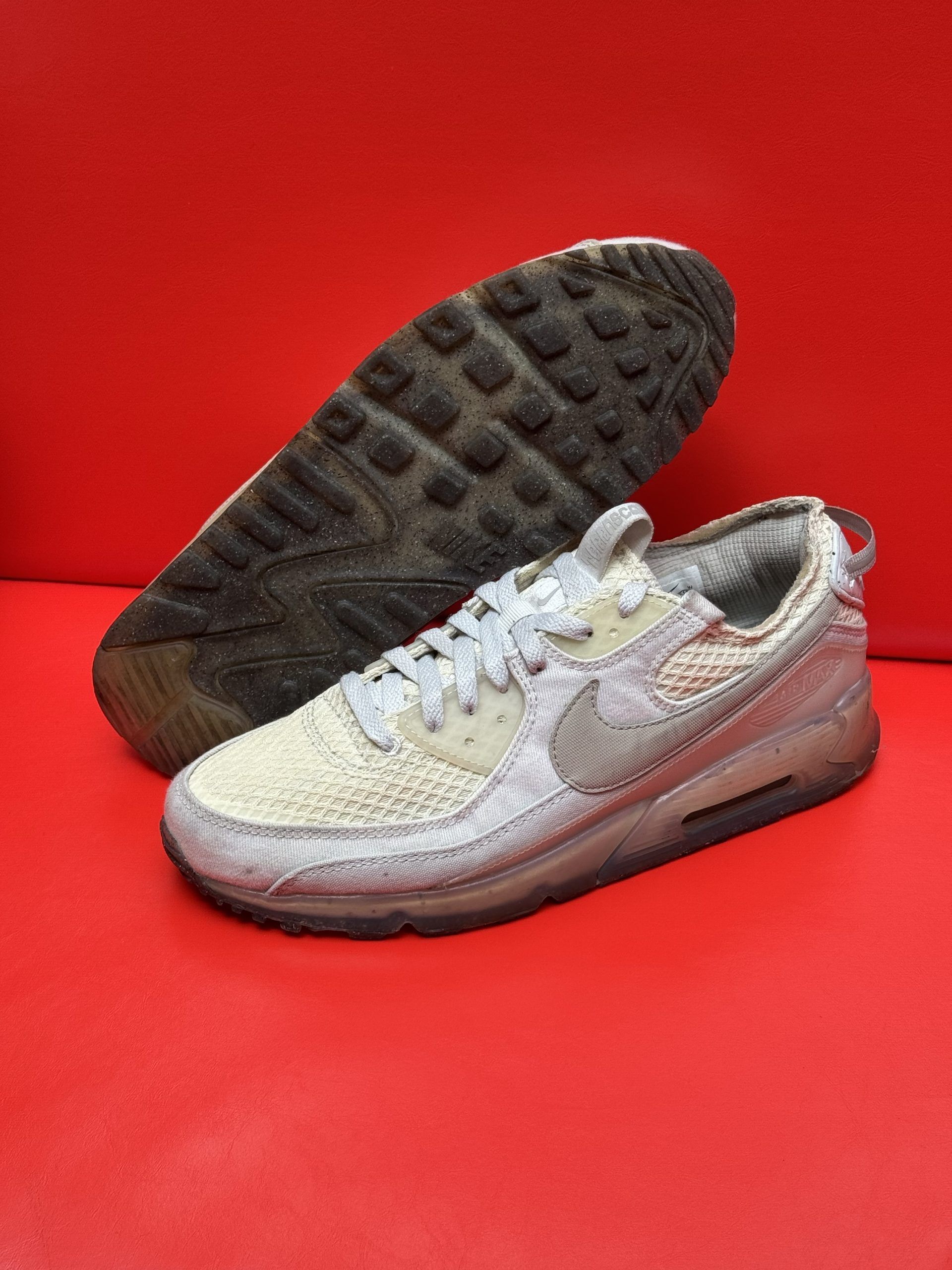 Light-colored Nike Air Max 90 sneaker with gray accents and dirty sole against a red background.