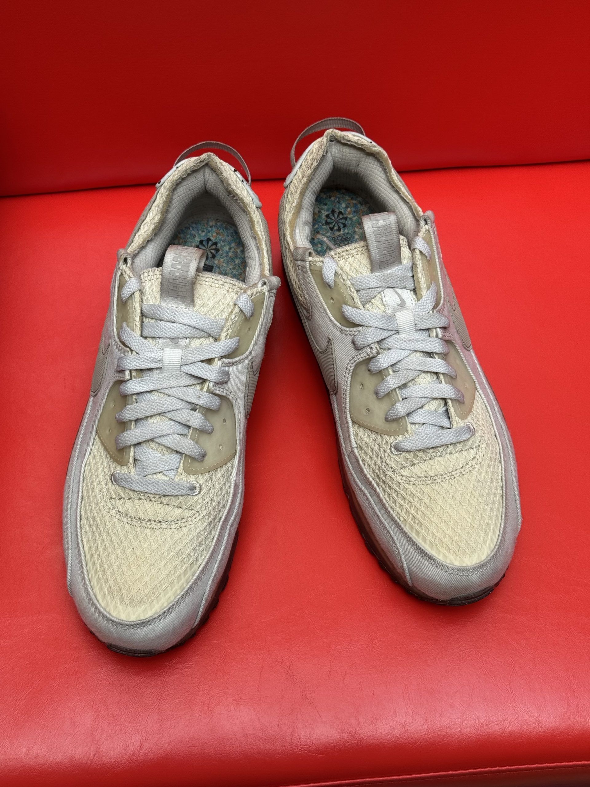 Light-colored Nike Air Max 90 sneaker with gray accents and dirty sole against a red background.