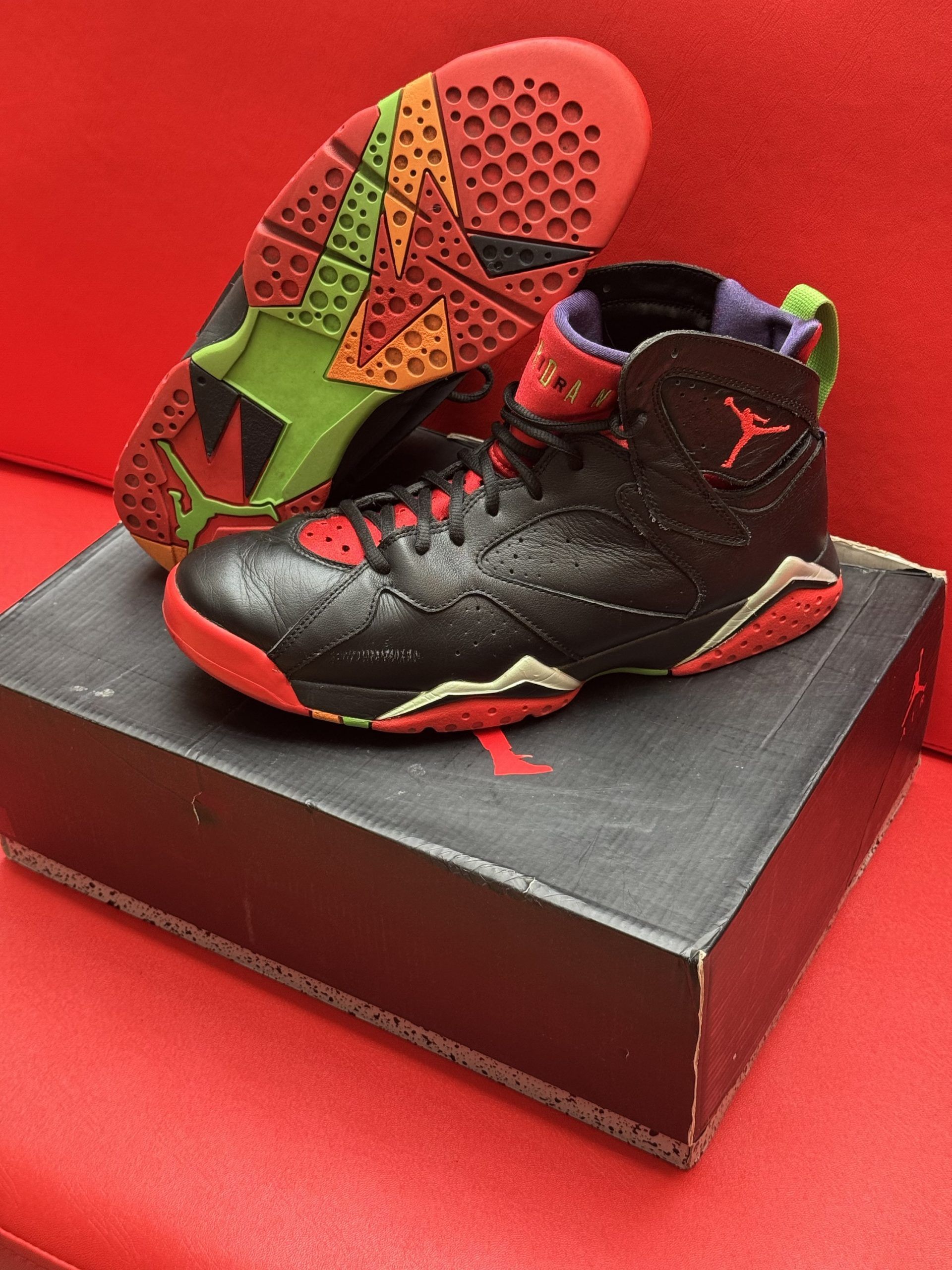 Black, red, and gold Air Jordan 7 sneakers on top of a black shoebox with a colorful sole on a red surface.