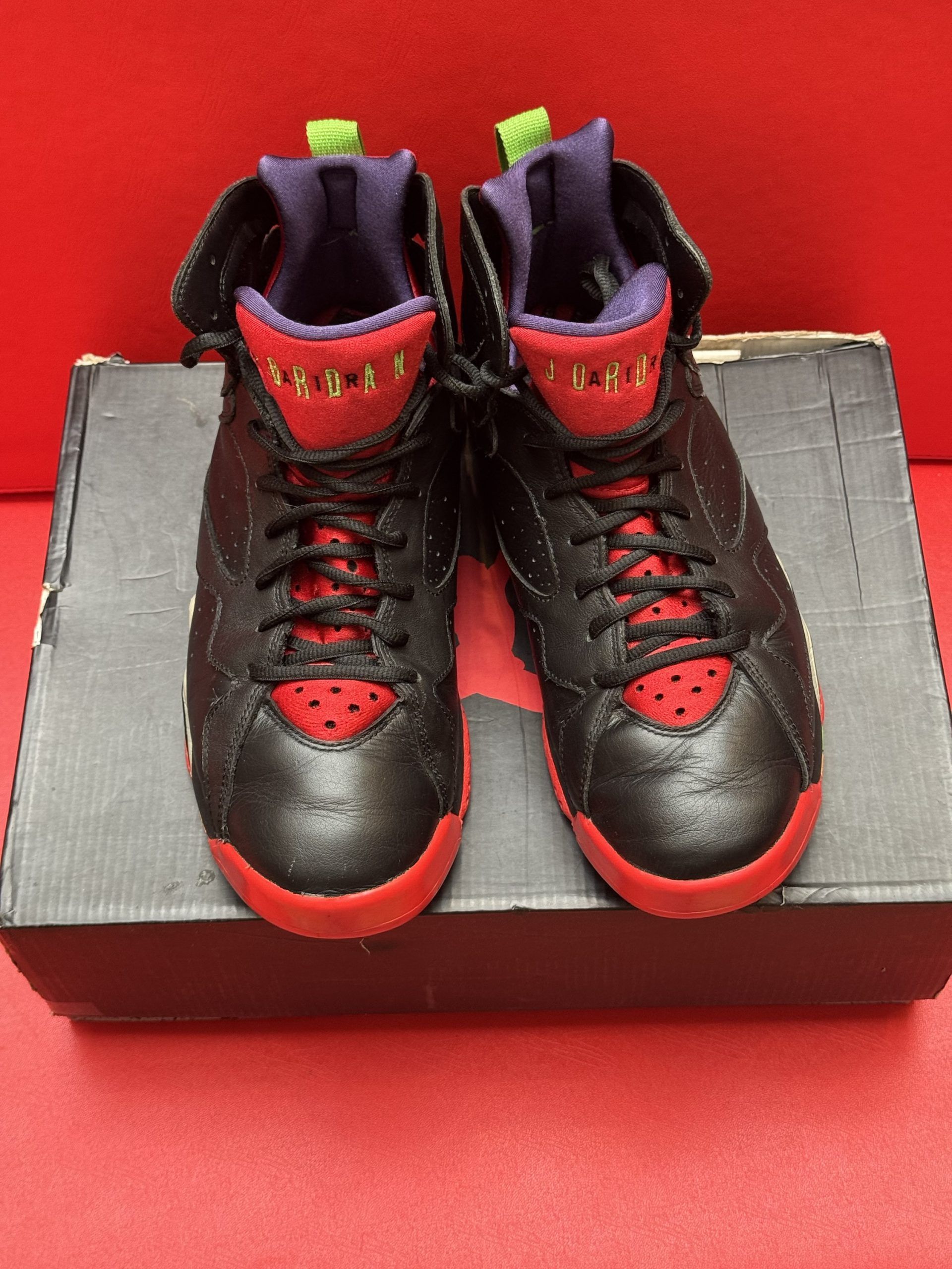 Black, red, and gold Air Jordan 7 sneakers on top of a black shoebox with a colorful sole on a red surface.