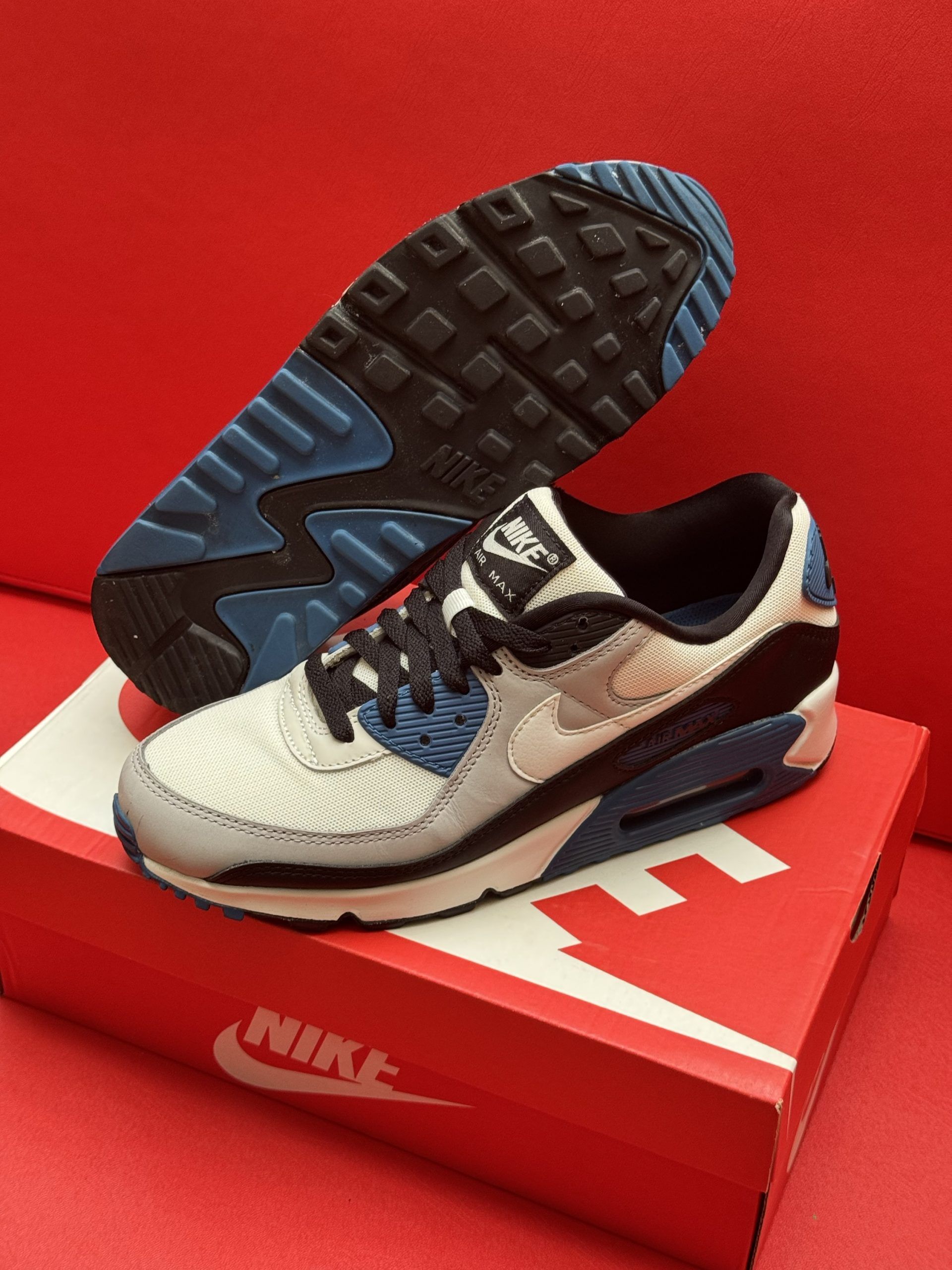 Nike Air Max 90 sneakers on a red box. One shoe on the box, the other shoe's sole is visible. White, blue, and black colors.