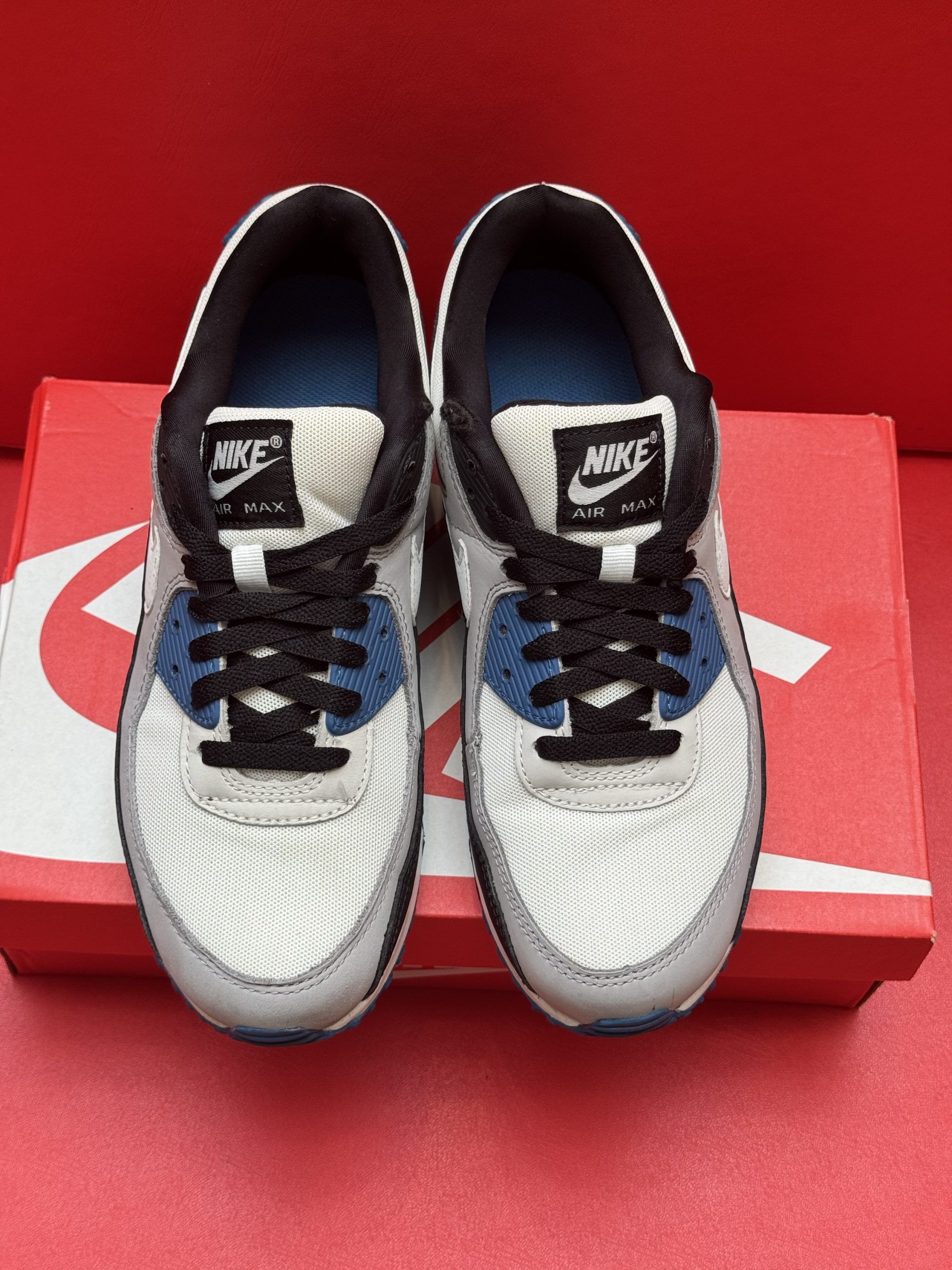 Nike Air Max 90 sneakers on a red box. One shoe on the box, the other shoe's sole is visible. White, blue, and black colors.
