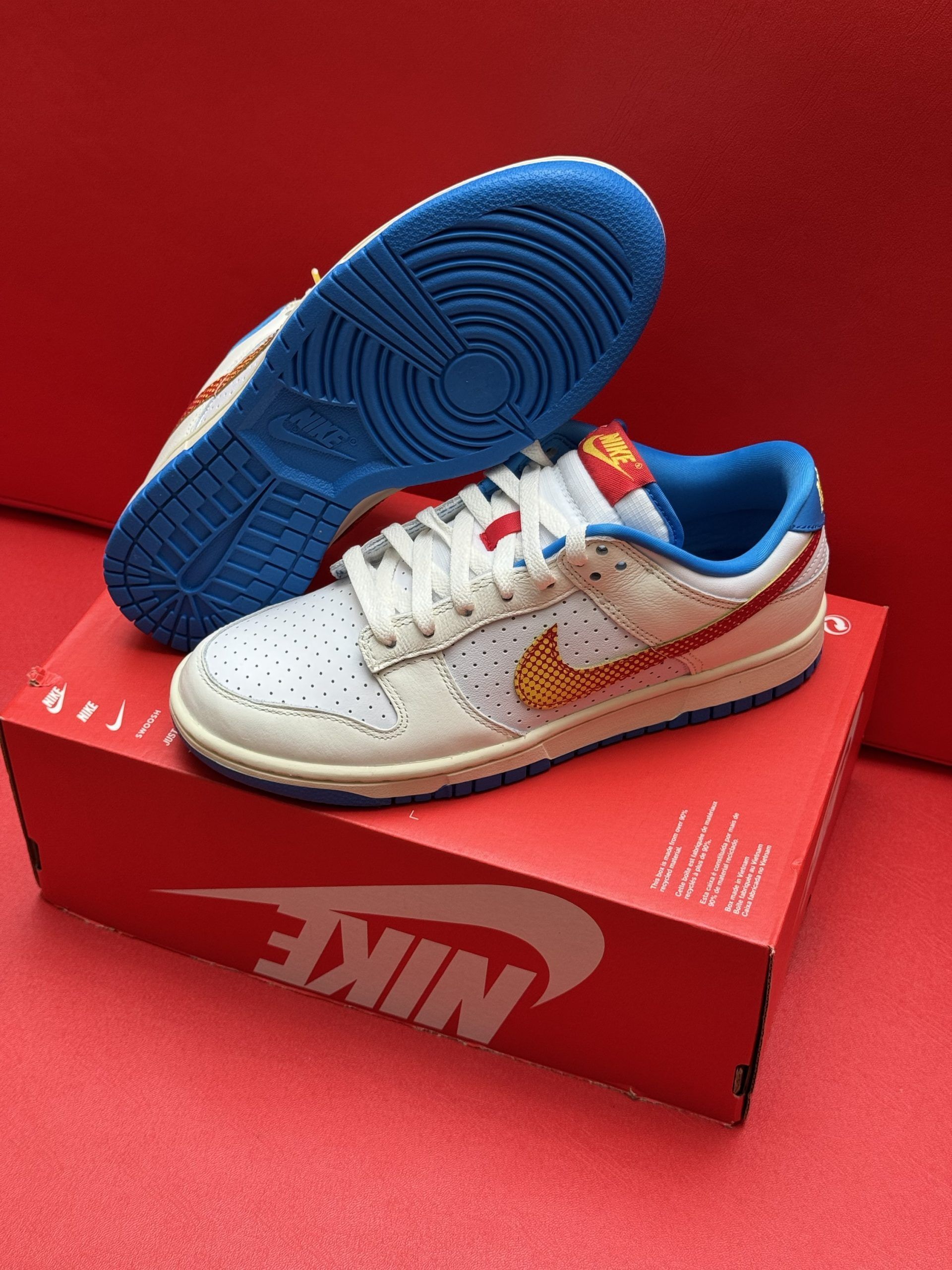 White Nike Dunk low sneakers with yellow and red accents on a red box, blue sole.