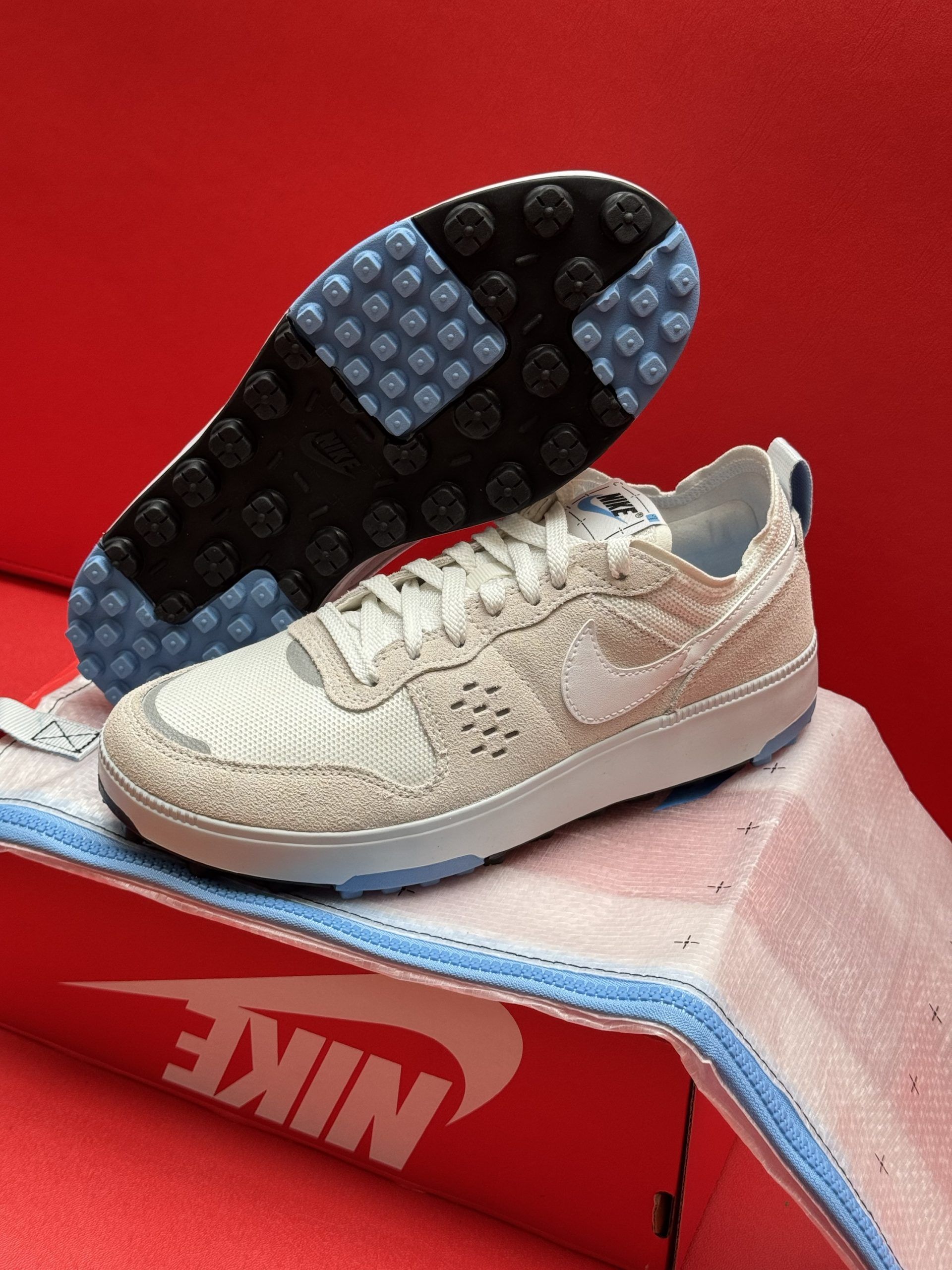 White Nike sneakers with a blue sole and logo, resting on a Nike box, set against a red background.