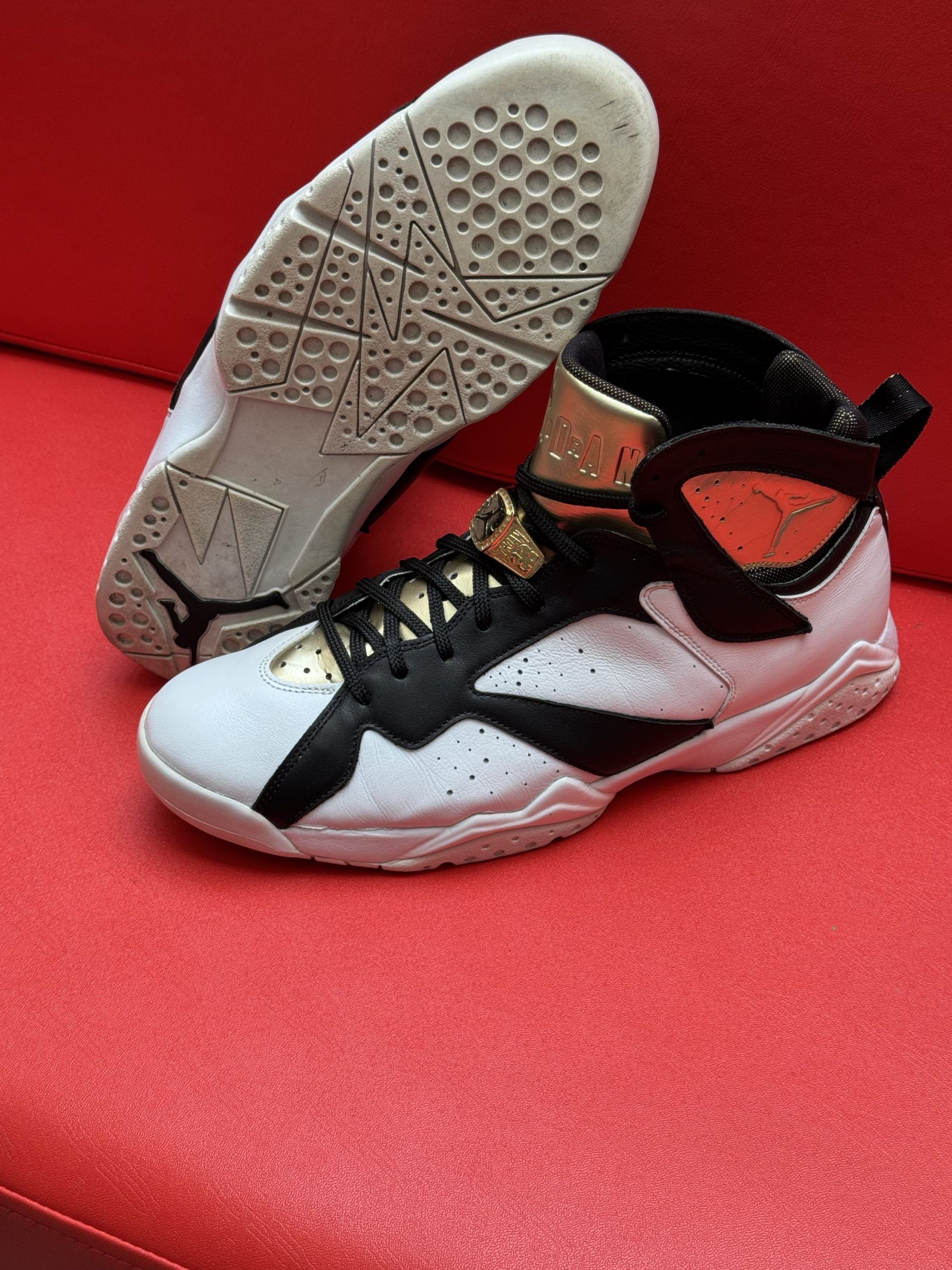 White, black, and gold Air Jordan 7 sneakers on a red surface.