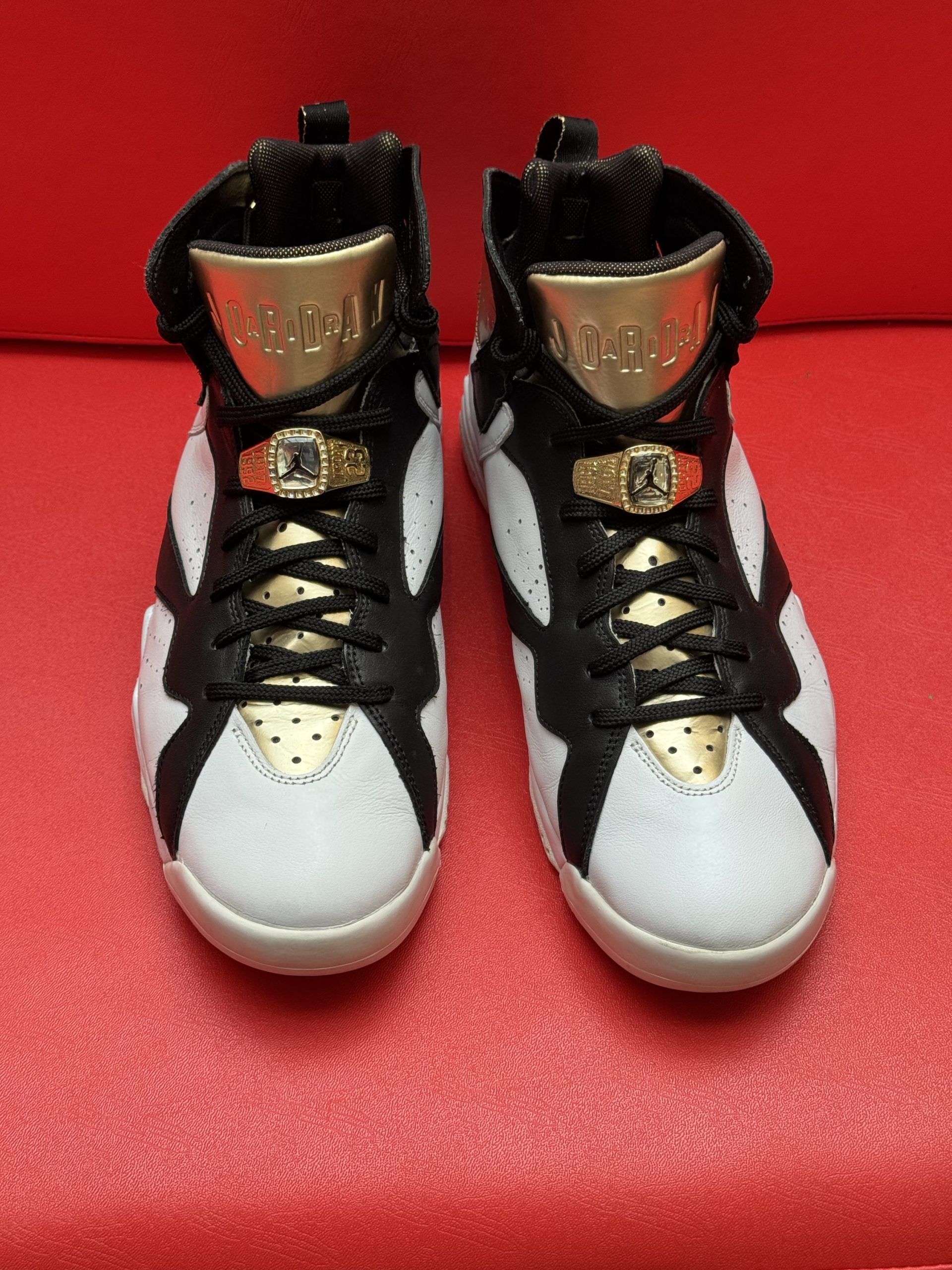 White, black, and gold Air Jordan 7 sneakers on a red surface.