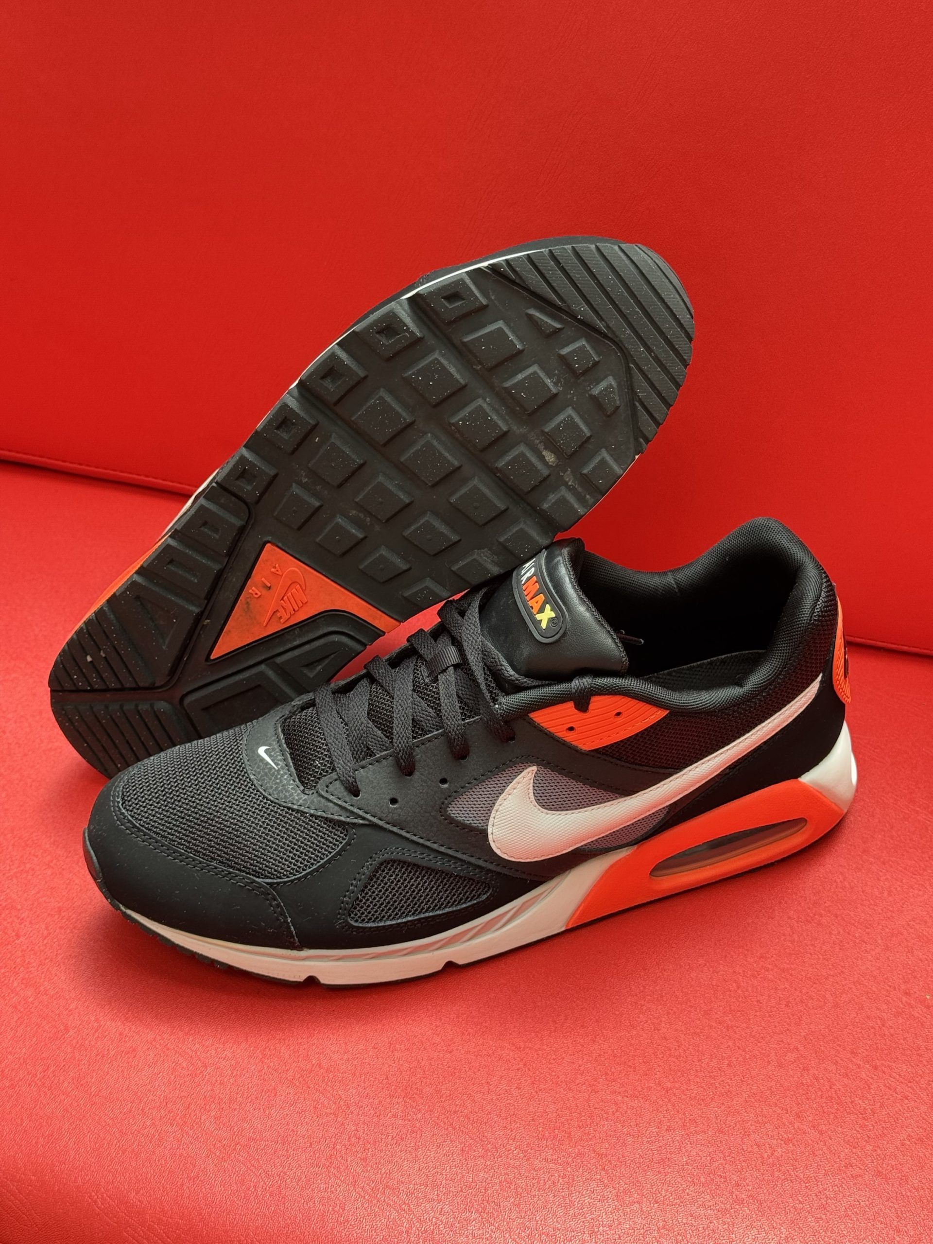 Black and orange Nike Air Max sneakers on a red surface. One shoe is upside down, revealing the sole.