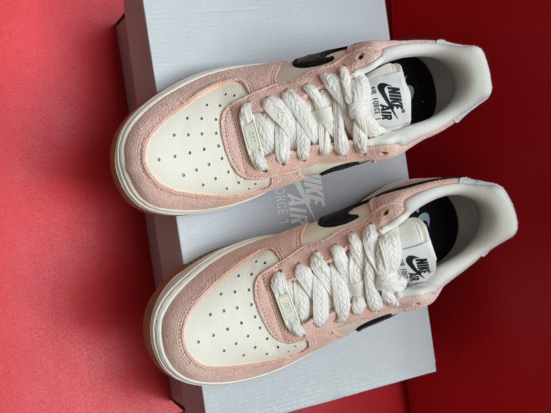 Pink and cream Nike Air Force 1 sneakers with black swoosh, sitting on white box, with the sole visible.
