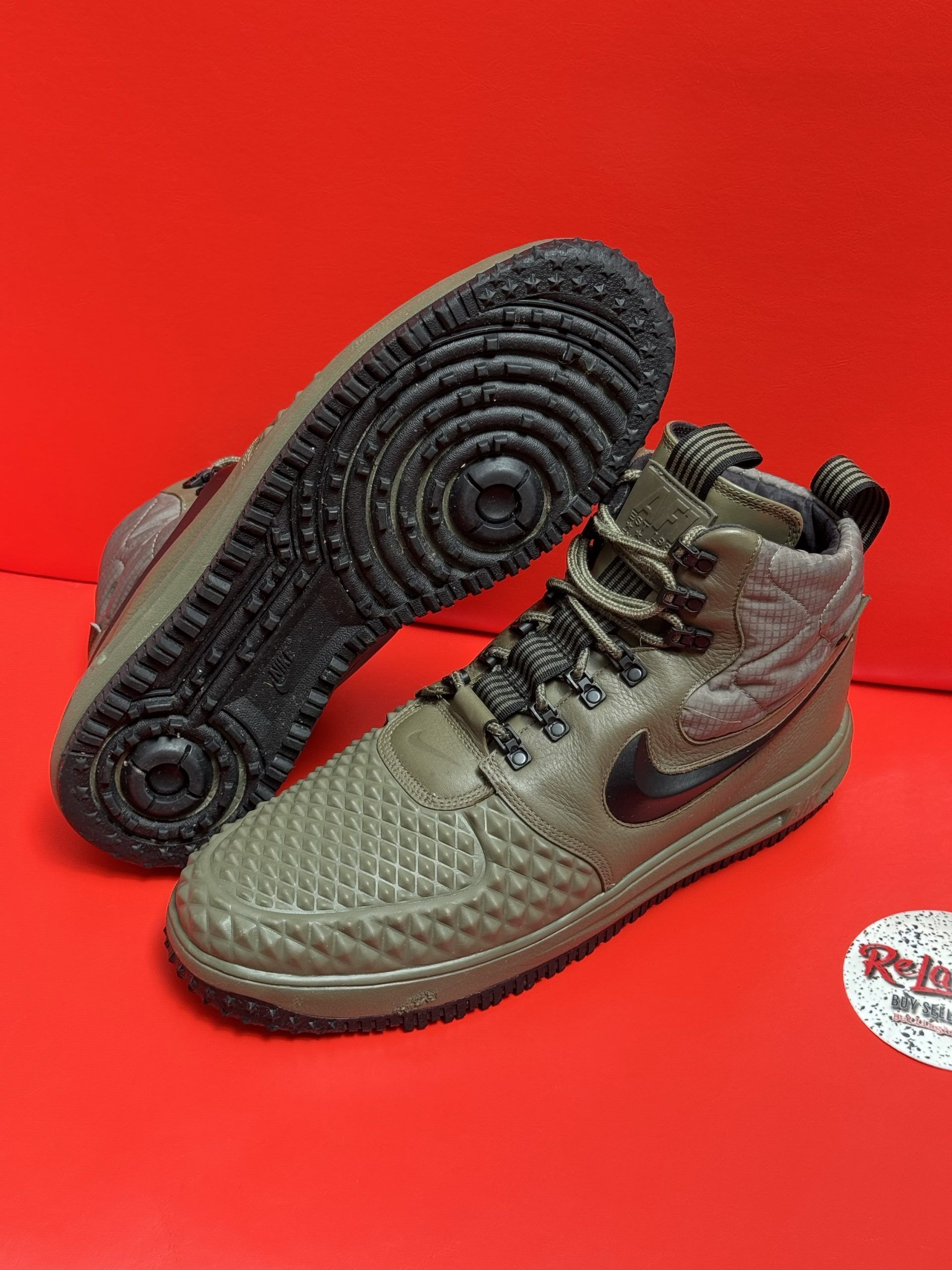Olive green Nike high-top sneakers with black soles, laces, and swoosh logo, on a red surface.