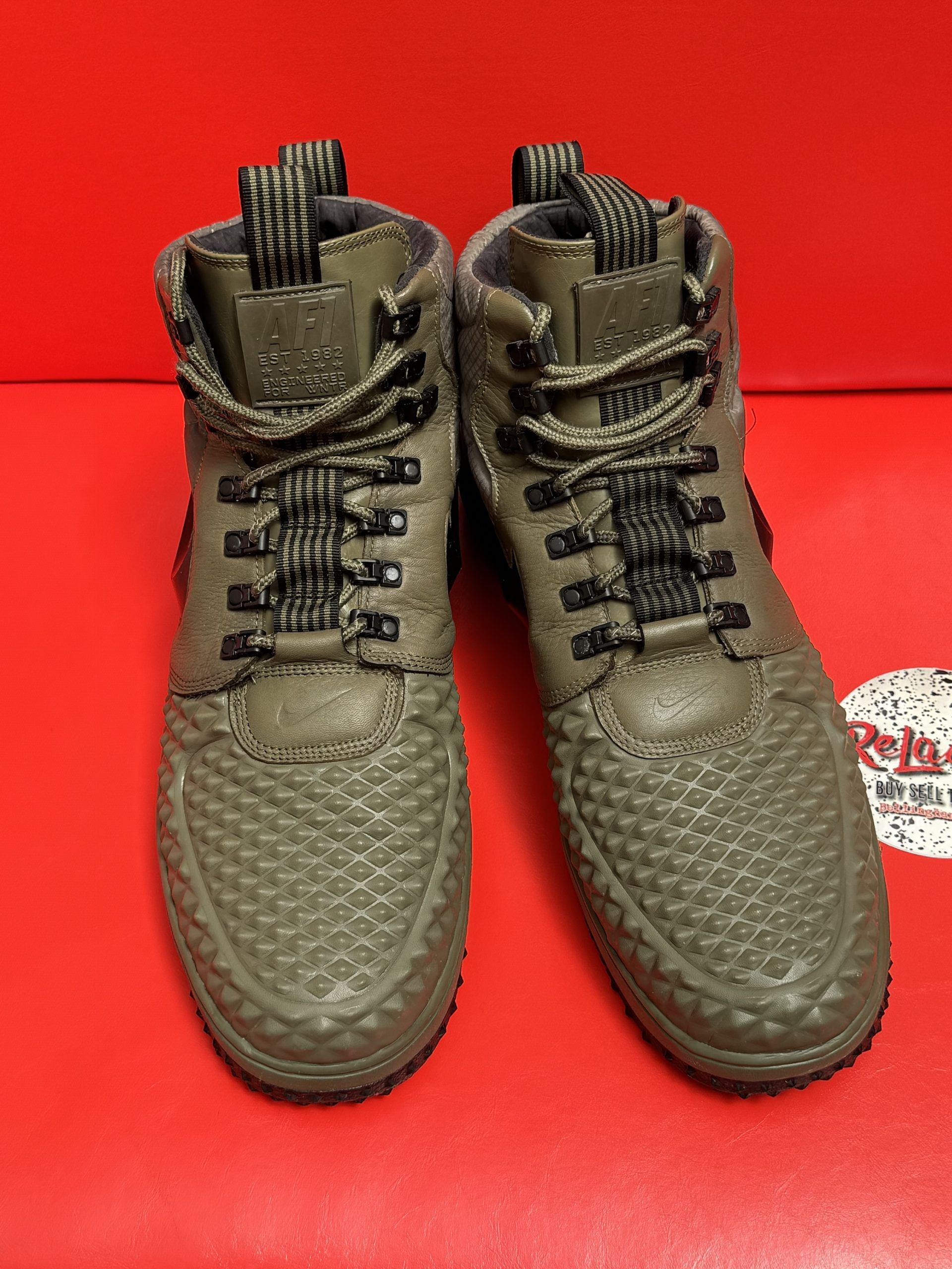 Olive green Nike high-top sneakers with black soles, laces, and swoosh logo, on a red surface.