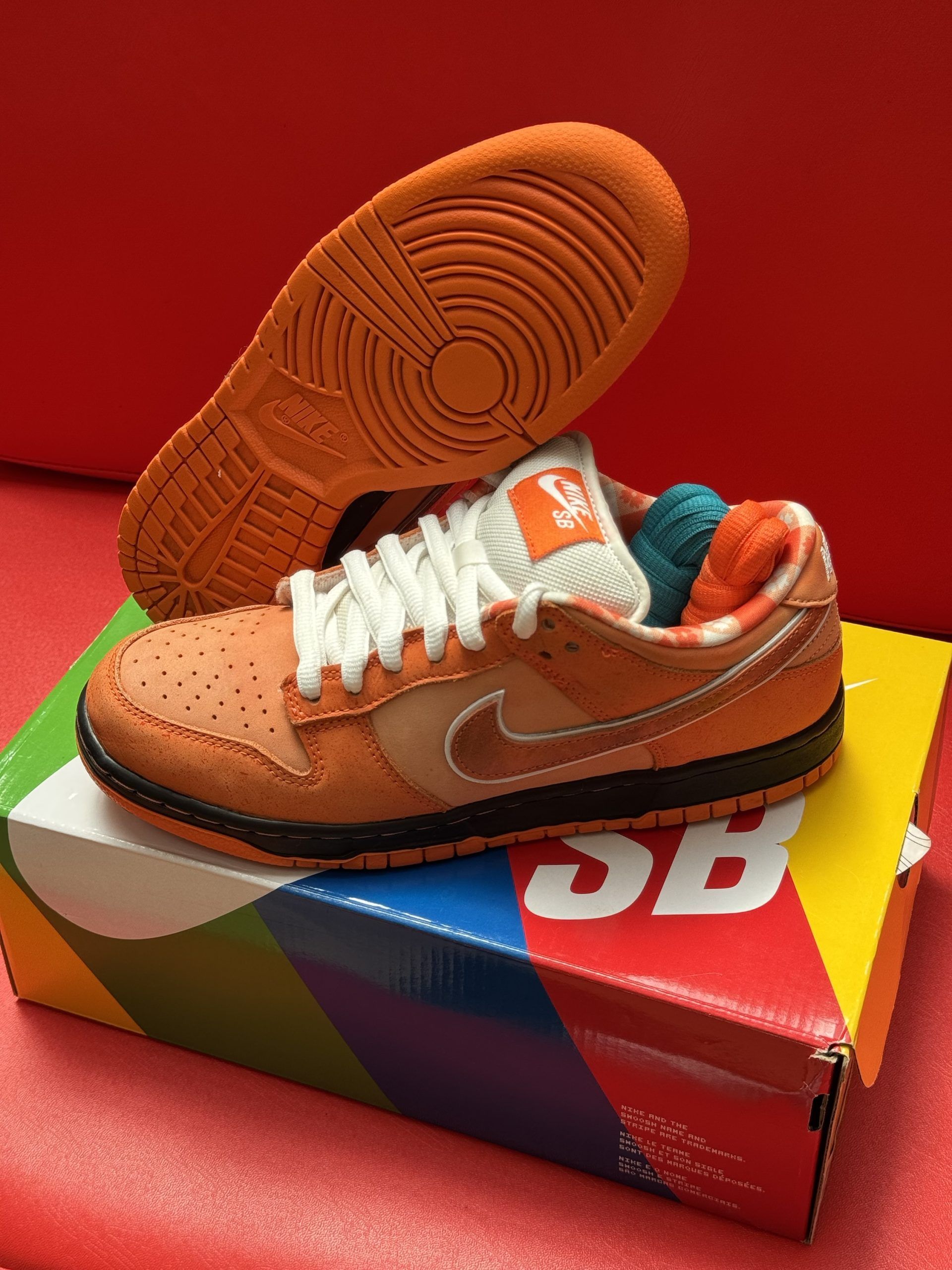 Orange Nike SB Dunk Low sneakers with white laces, sitting on a colorful shoebox, one sole up.