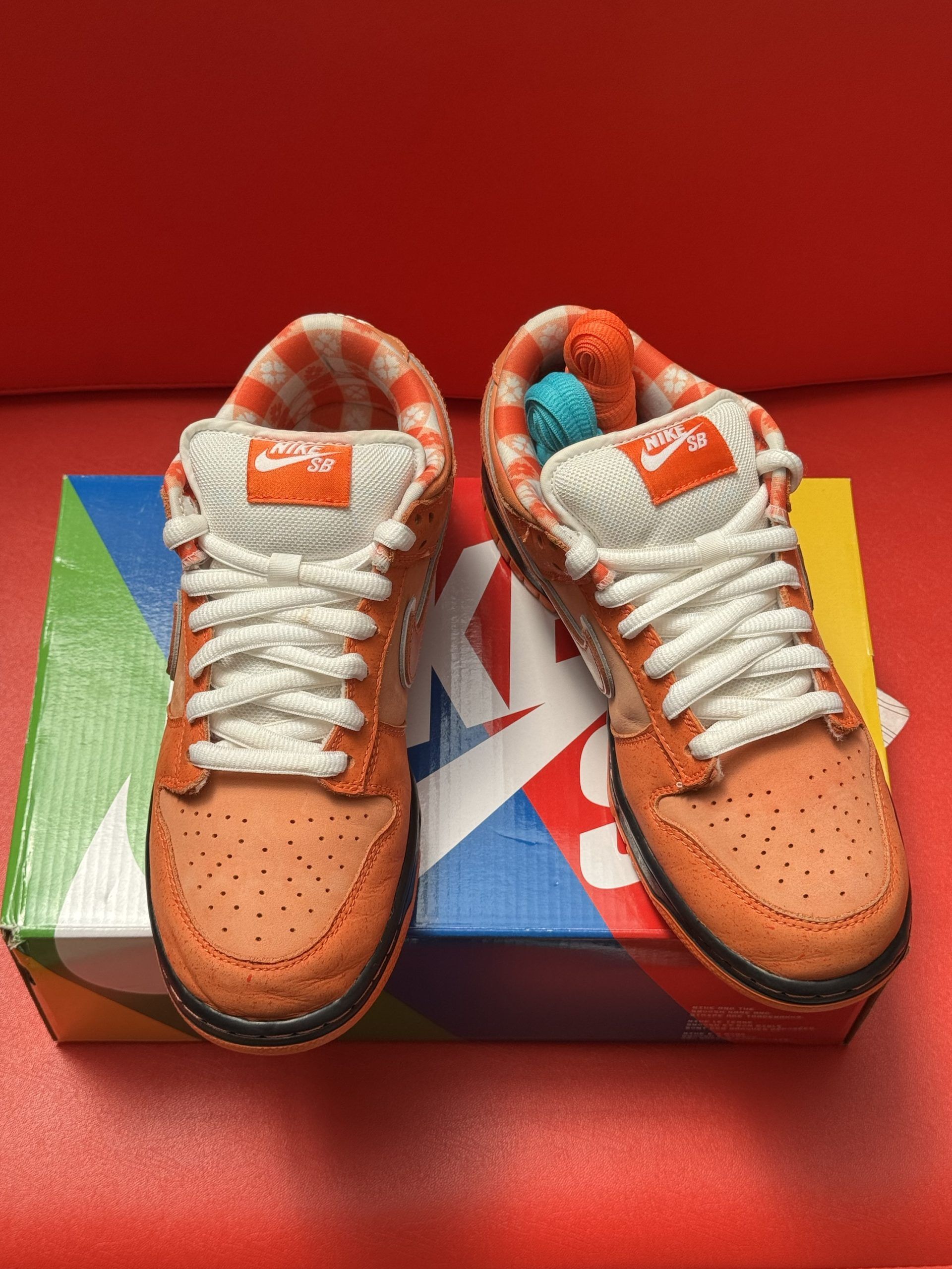 Orange Nike SB Dunk Low sneakers with white laces, sitting on a colorful shoebox, one sole up.