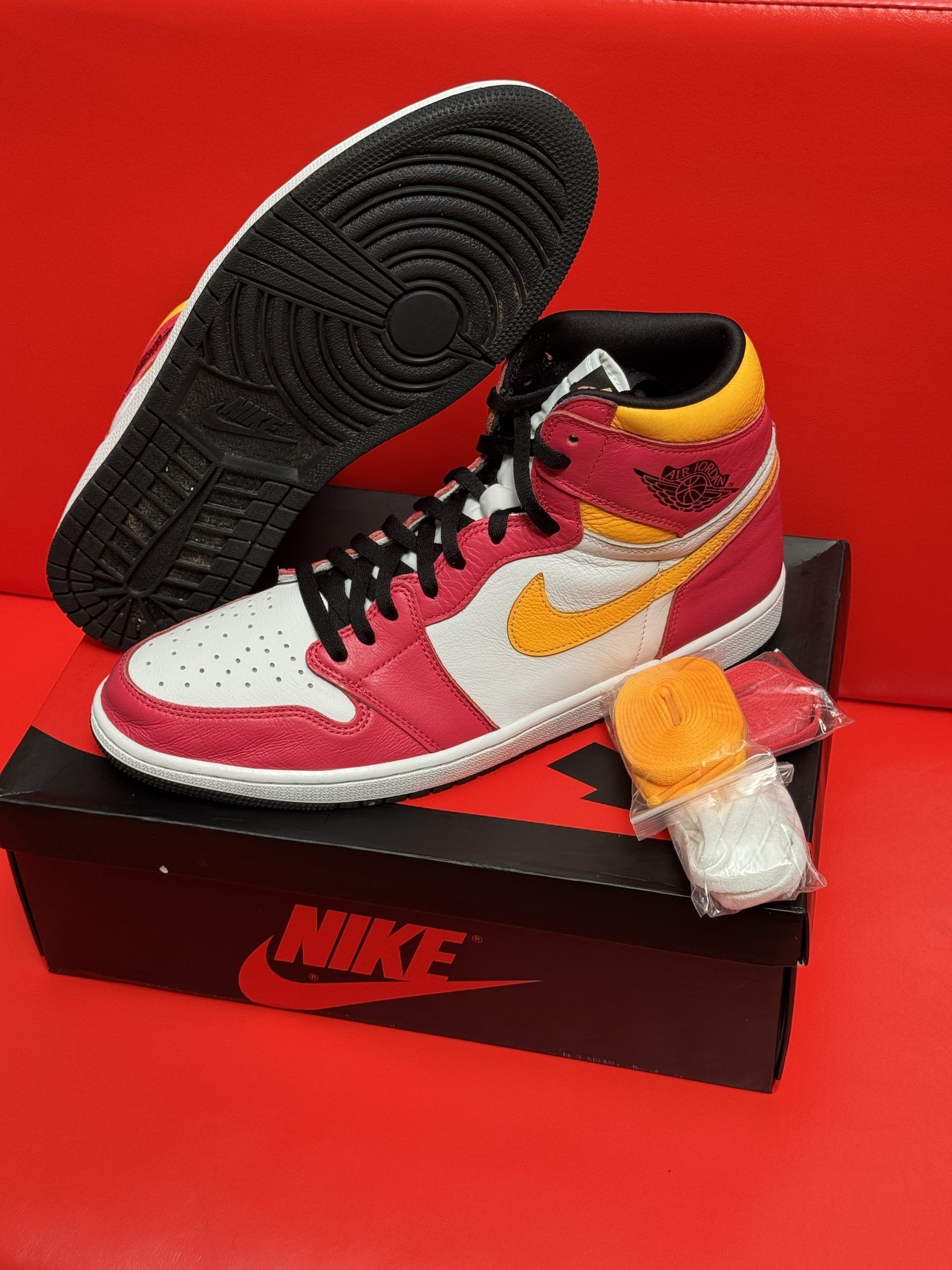Pink, yellow, and white Air Jordan 1 high-top sneaker on black Nike box, extra orange laces, black sole, red background.