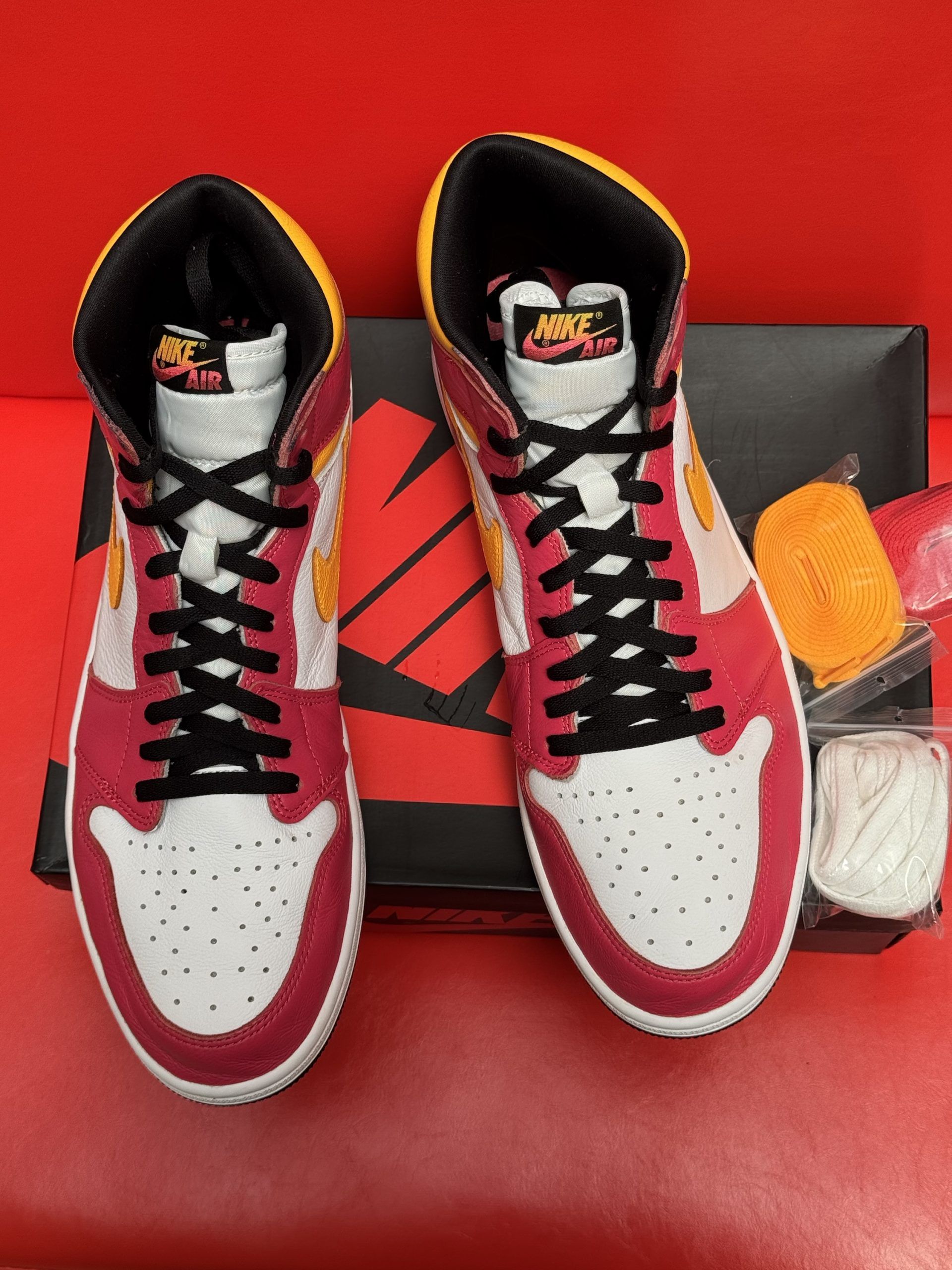 Pink, yellow, and white Air Jordan 1 high-top sneaker on black Nike box, extra orange laces, black sole, red background.