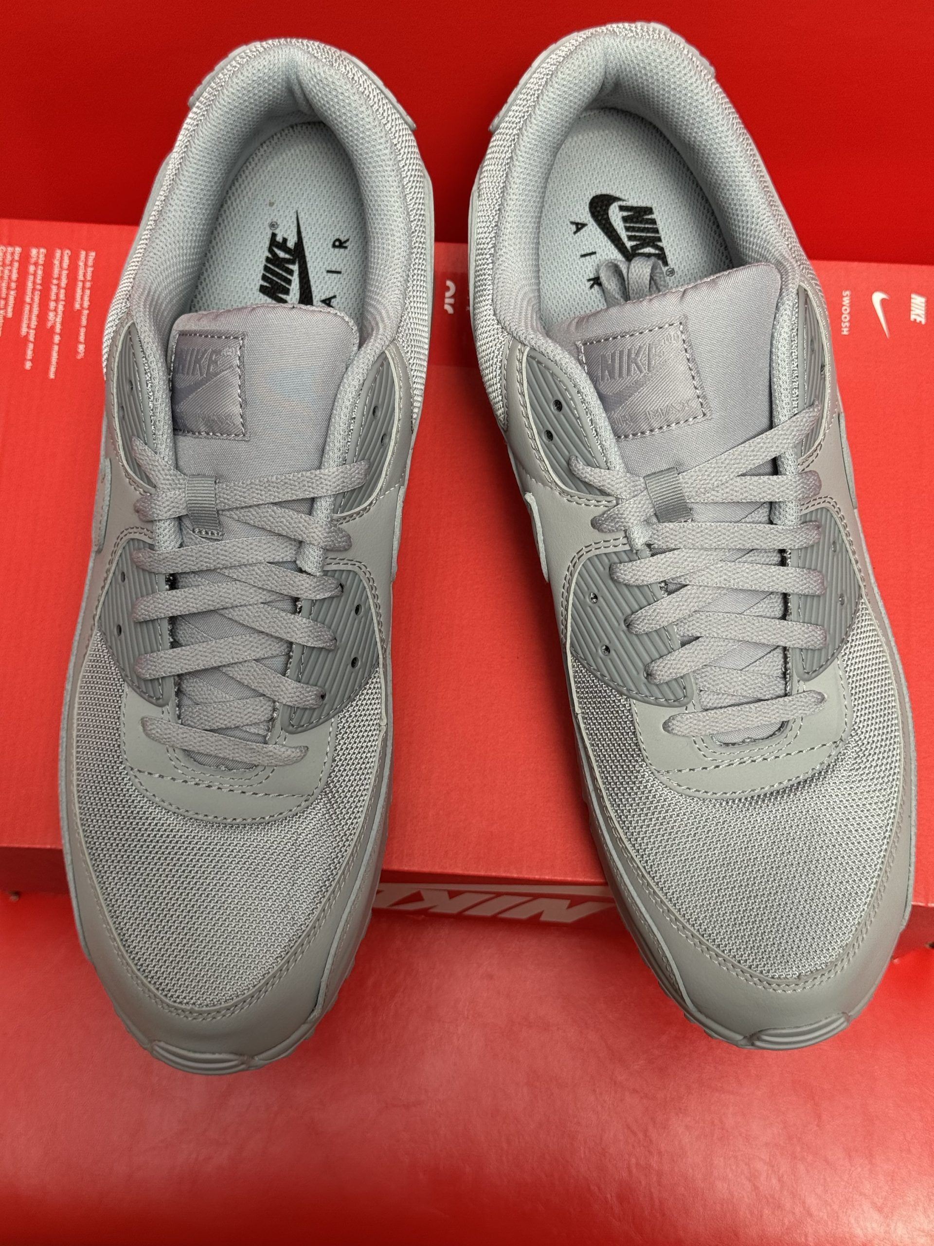 Gray Nike Air Max 90 sneakers on a red Nike box, with the sole visible. Red background.