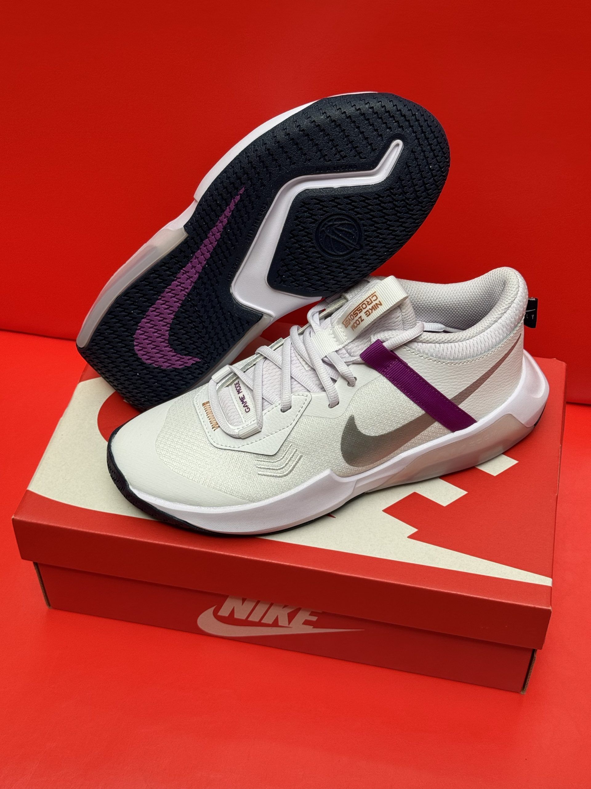 White Nike basketball sneakers with purple accents, on a Nike box, set against a red background.
