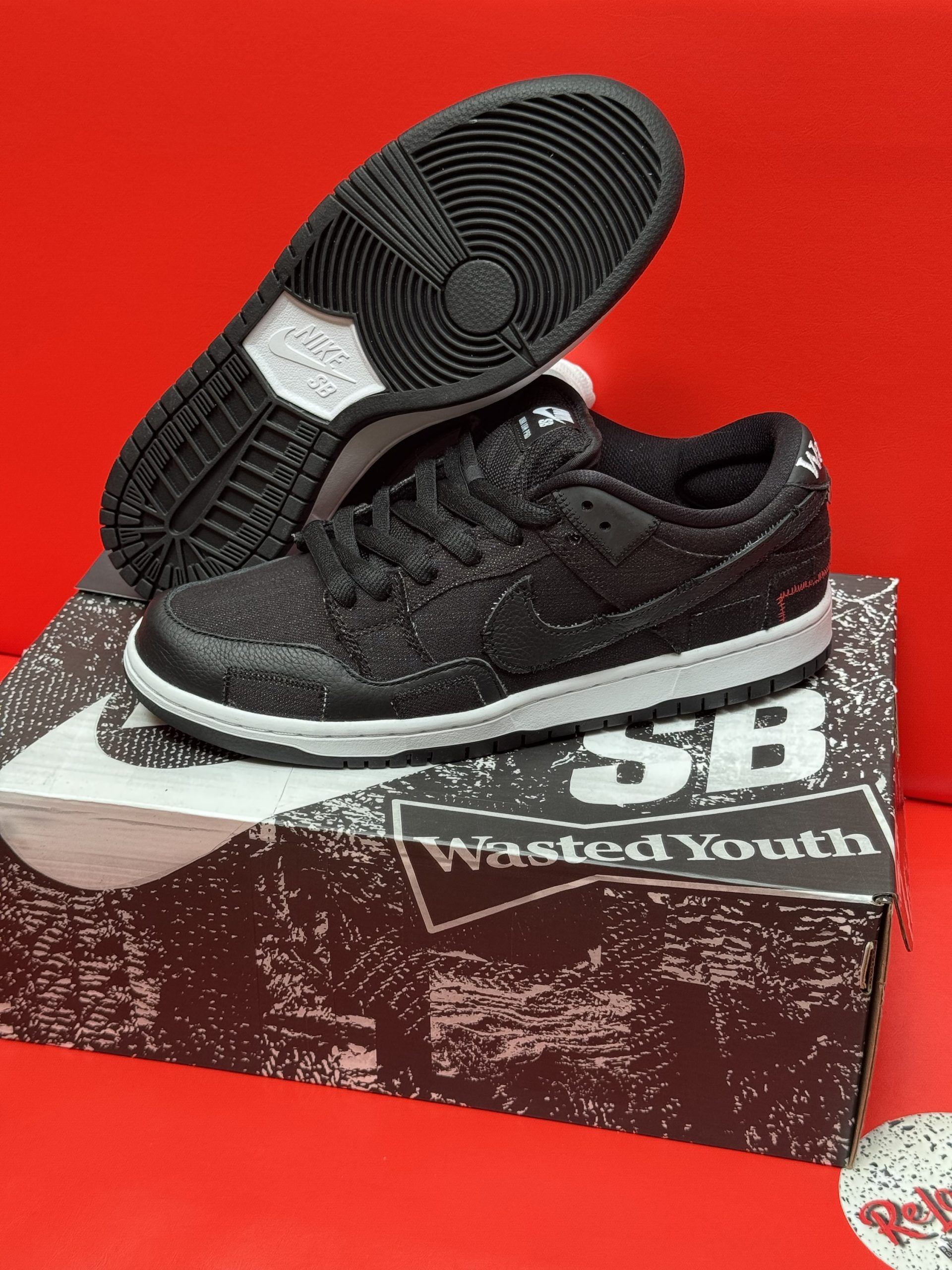 Black Nike SB Dunk Low sneakers with white soles, sitting on a branded box with a red background.