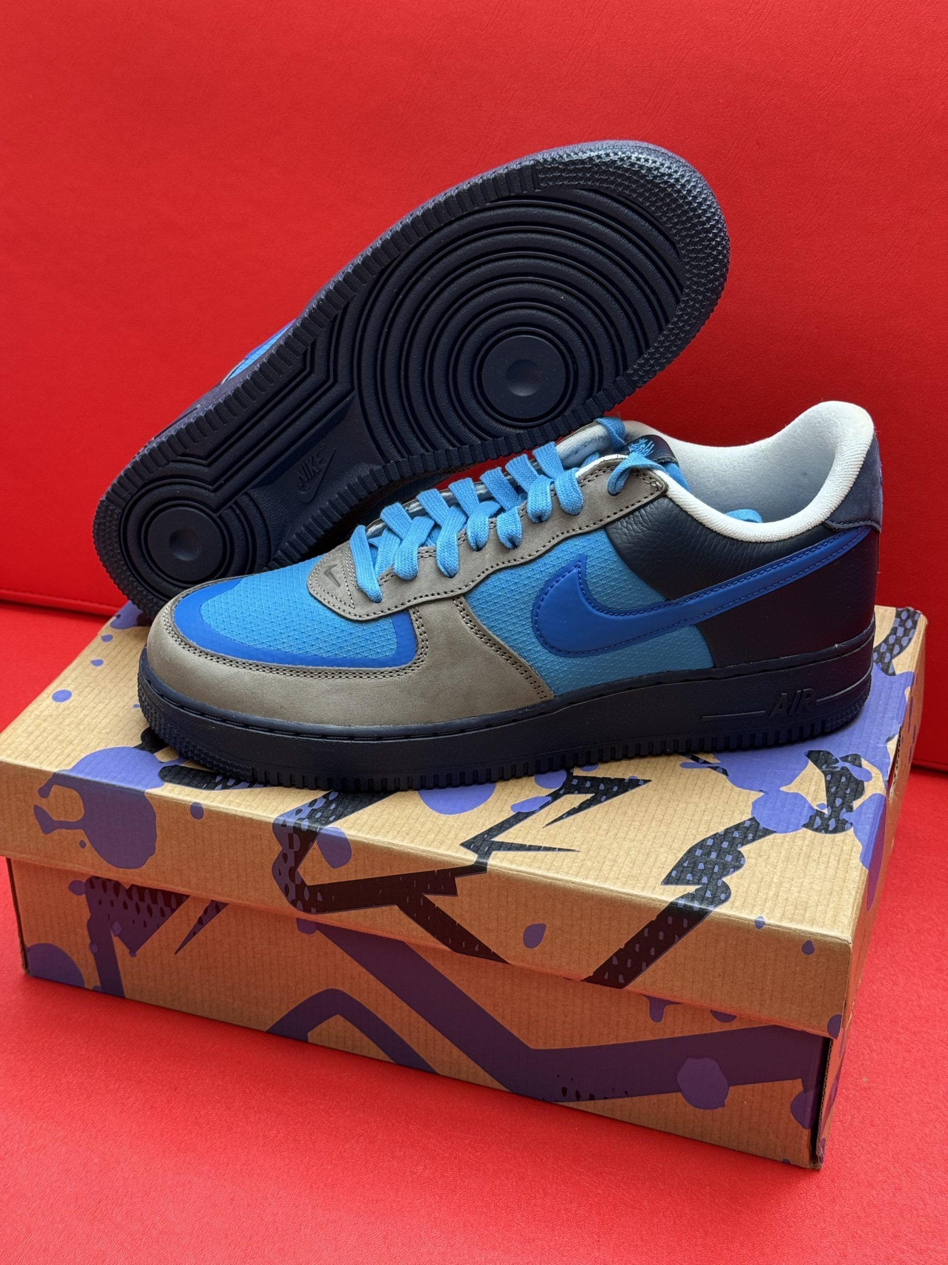Blue and gray Nike Air Force 1 sneakers on splatter-painted box, against a red background.
