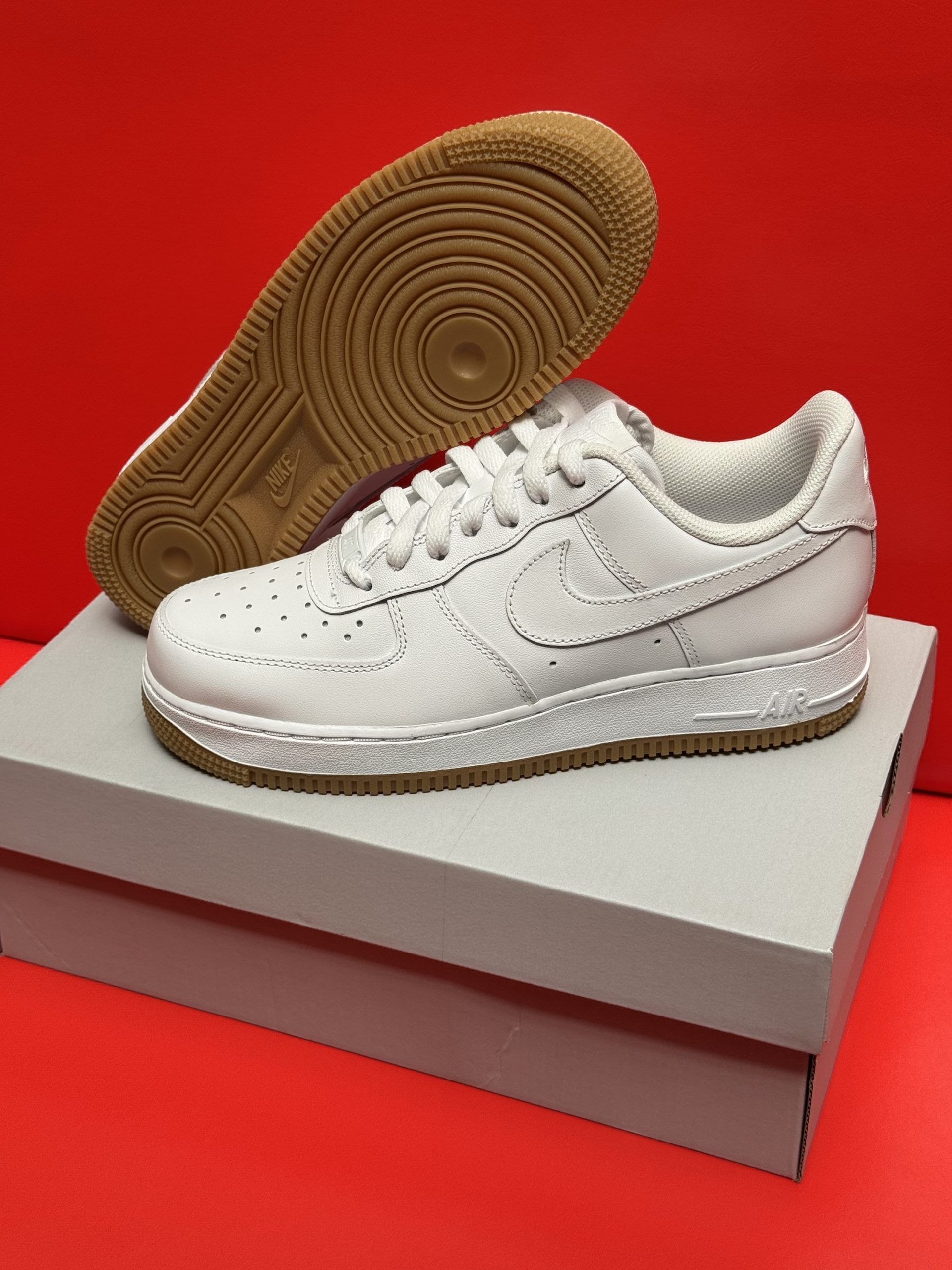White Nike Air Force 1 sneakers with brown soles on a gray box, red background.