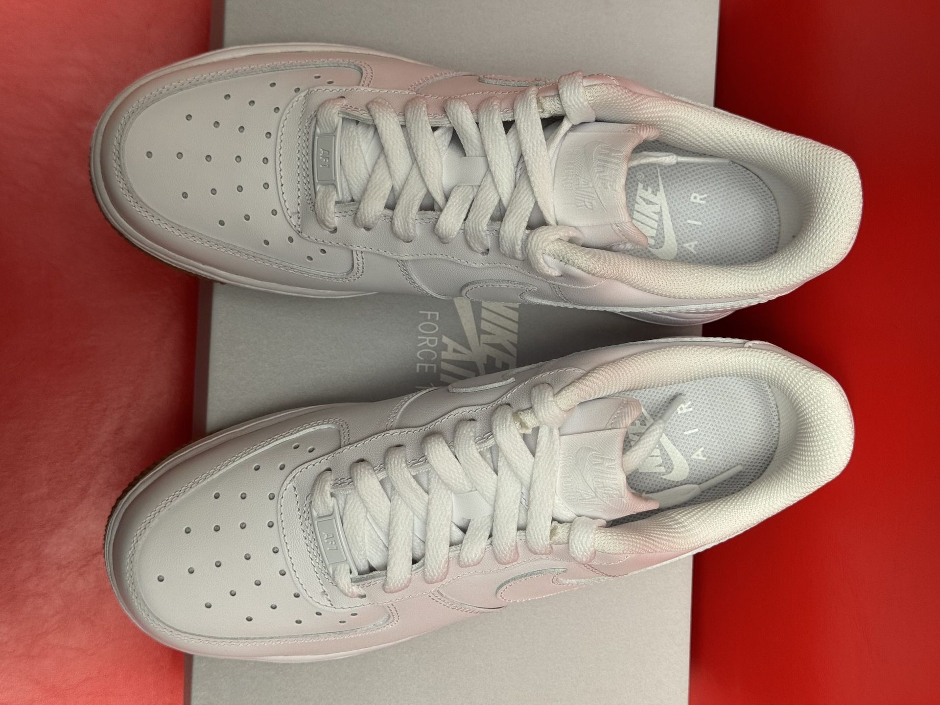 White Nike Air Force 1 sneakers with brown soles on a gray box, red background.