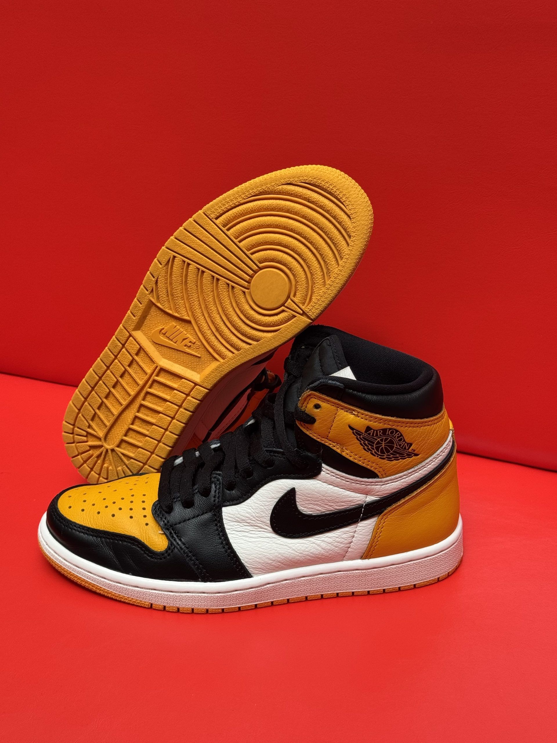 Yellow, black, and white high-top Air Jordan 1 sneaker with a visible yellow sole against a red backdrop.