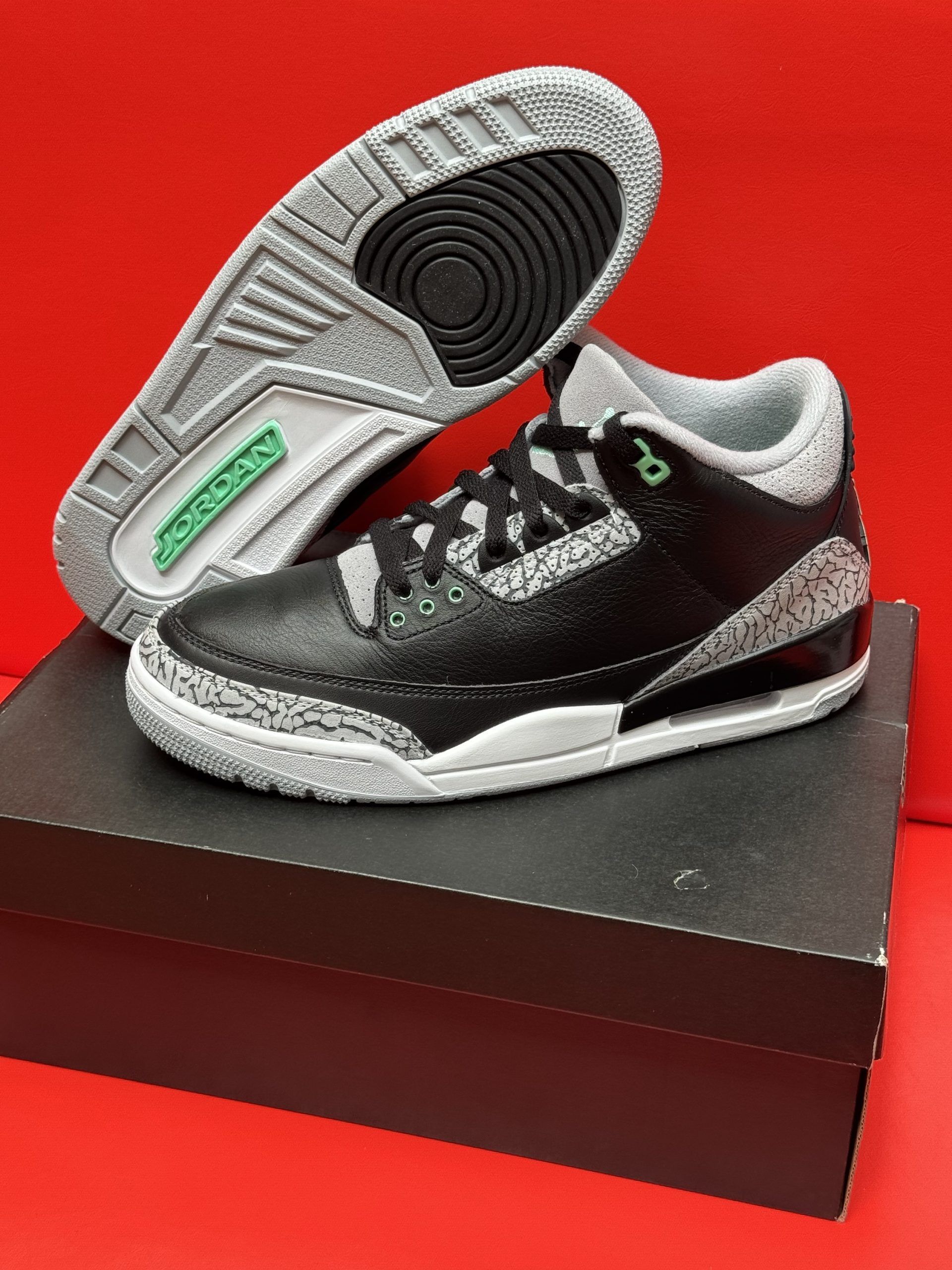 Black and gray Air Jordan 3 sneakers on a black box, one shoe upside down showing its sole; red background.